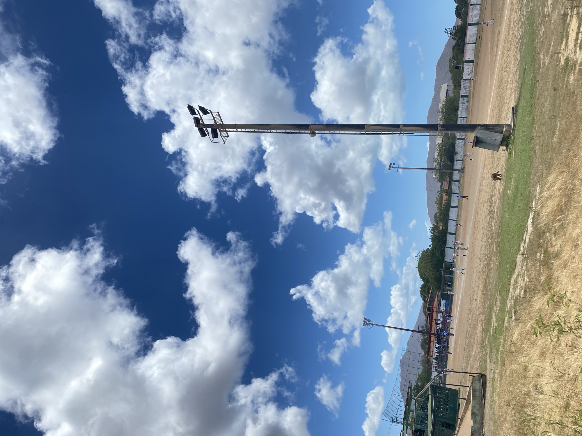 The image shows a baseball field in Juan Griego, Venezuela. The sky is blue with scattered white clouds. A tall metal light pole stands near the field. In the background, mountains are visible. Several people are on the field playing baseball. Fences surround the field, and there is a dugout in the foreground. The field is dry, with some grass.