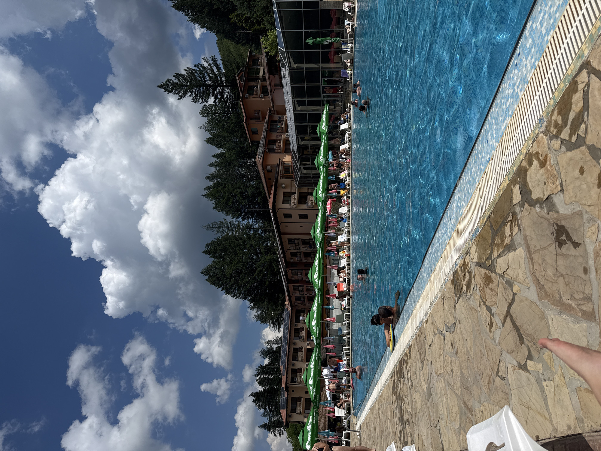 This image depicts an outdoor swimming pool area at a resort in Chiflik, Bulgaria. The pool is filled with clear blue water and is surrounded by a stone patio. A few people are in the water, swimming or relaxing. Around the pool, lounge chairs and green umbrellas are set up, with some people sitting and sunbathing. In the background, there is a multi-story hotel with a facade of brown and beige tones, featuring balconies and solar panels on the roof. Adjacent to the hotel is a glass-enclosed structure. The area is bordered by a dense forest of pine trees, suggesting a mountain or hilly terrain. The sky is blue with scattered white clouds.