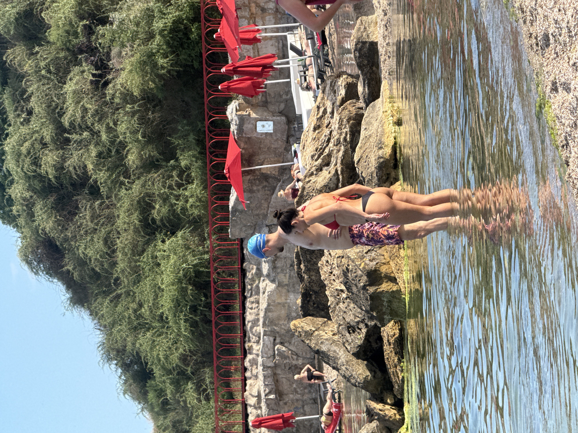The image shows a group of people at a rocky beach with clear water and lush green vegetation on a hill in the background. A couple, a man wearing a blue swimming cap and patterned swim trunks, and a woman in a red bikini, are standing in the shallow water. In the background, several beach umbrellas are set up, and people are relaxing on lounge chairs on a higher rocky terrace. The scene appears to be a beach resort or a natural coastal area. The presence of lush vegetation suggests a temperate or Mediterranean climate. The overall atmosphere is relaxed and summery. It is not possible to determine the exact location in Odesa, Ukraine from the image alone, but it is consistent with coastal areas in the region.