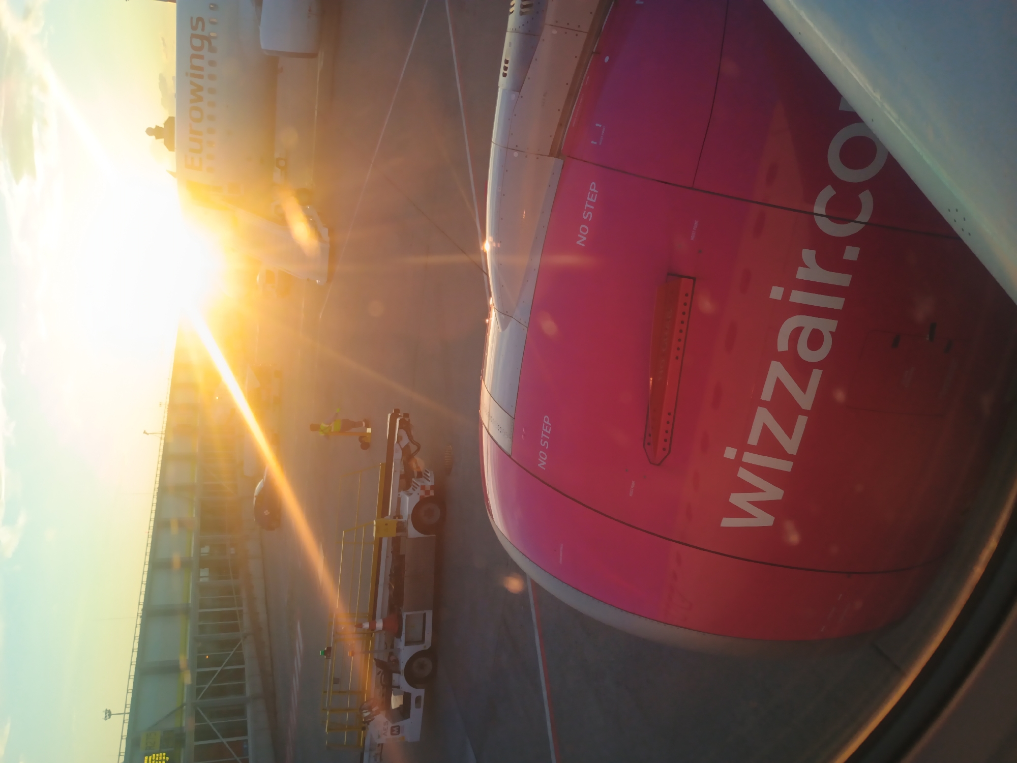 This is an image taken from inside an airplane window, looking out at an airport tarmac during sunset. The sun is setting, casting a warm, golden light across the scene, with lens flares visible. In the foreground, the pink engine cowling of the airplane is visible, with the Wizz Air logo prominently displayed. The text "NO STEP" is also visible on the engine. In the background, another airplane with "Eurowings" written on its side is parked. An airport terminal building with large windows and a walkway can be seen to the left, and various ground support vehicles are present on the tarmac. A person wearing a high-visibility vest is seen standing near a vehicle. The scene is likely at Budapest Ferenc Liszt International Airport (BUD), given the presence of Wizz Air, a Hungarian airline.