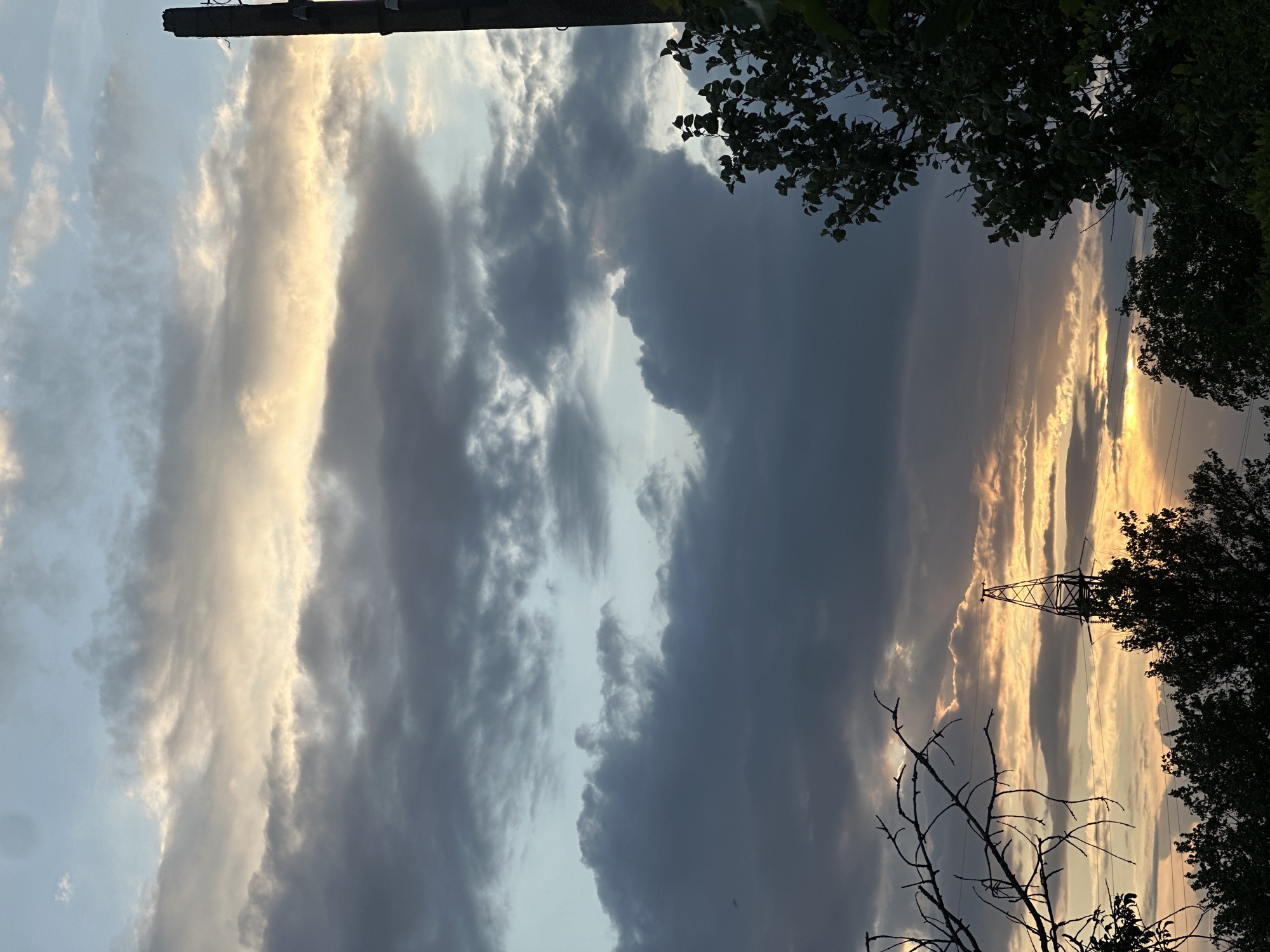 A dark, overcast sky with streaks of yellow and orange light breaking through the clouds, suggesting either sunrise or sunset. The foreground is dominated by the silhouettes of trees and branches against the sky. On the right side of the frame, a tall, dark, cylindrical object, possibly a utility pole, is visible. In the background, a power line tower stands silhouetted against the brightest part of the sky. The overall impression is a moody, atmospheric landscape.