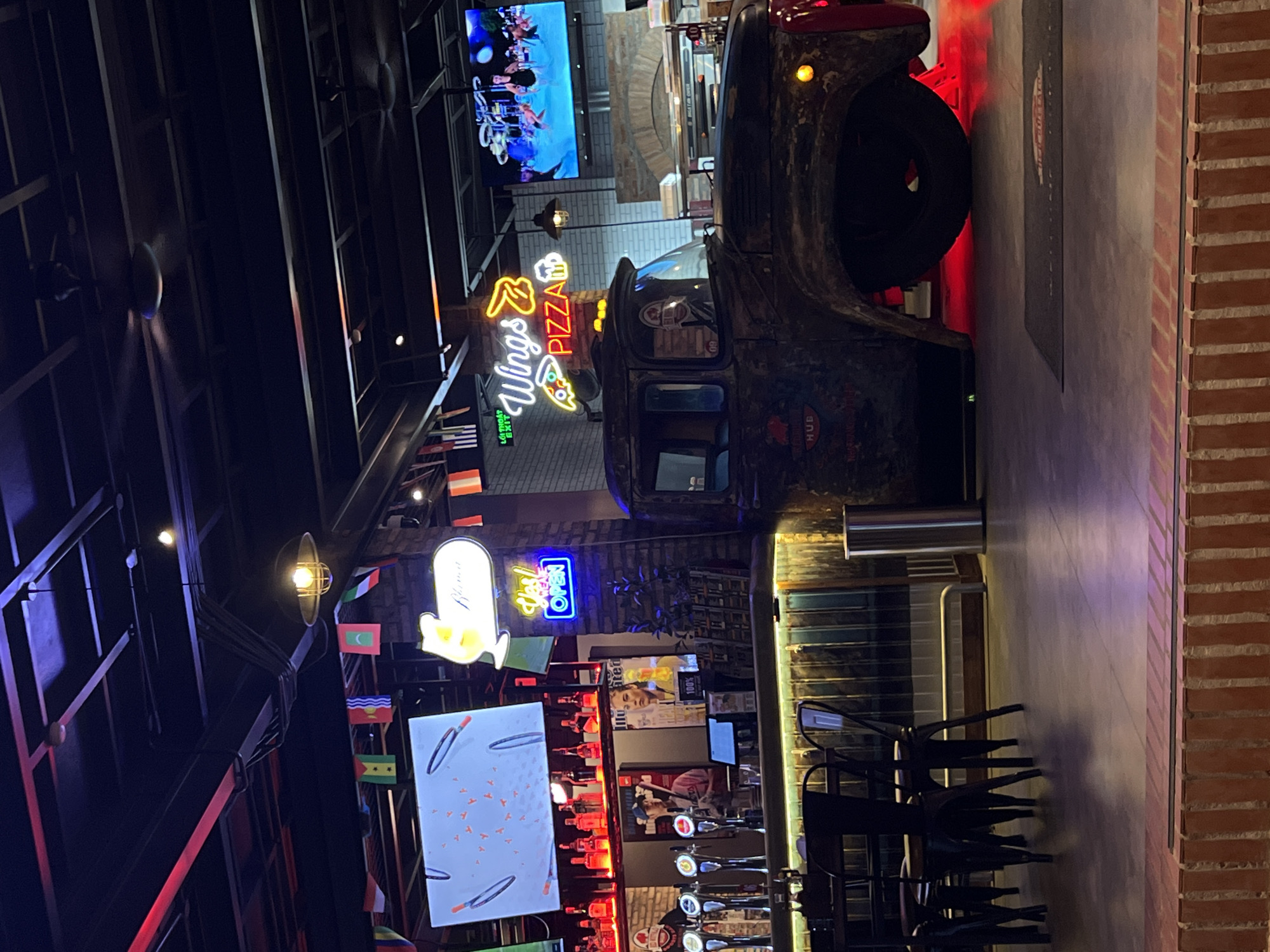 This indoor photograph, captured in Bac Nha Trang Ward, Vietnam, portrays a vibrant and dimly lit restaurant or bar environment, likely during the evening or night. The scene is characterized by a prominent, rustic, vintage-style truck, possibly serving as a decorative focal point or a food station. The truck exhibits a weathered appearance, featuring a yellow headlight and a red light accent at its rear. The word "HUB" is discernible on its side.
Above the truck, a bright neon sign glows, clearly spelling "Wings & Pizza" and incorporating icons of a chicken wing and a pizza slice. Adjacent to this, a blue neon "OPEN" sign and a white sign shaped like a beer mug, possibly reading "BIARITZ," contribute to the lively ambiance. A green sign to the left indicates "LỐI THOÁT EXIT" (Vietnamese for "Exit").
A large flat-screen television mounted on a brick wall displays a performance or concert, showing multiple individuals on a stage with musical instruments. Below this, a well-equipped bar area is visible, featuring numerous beer taps, bottles, and glassware. Stacked black chairs are arranged nearby. Another screen on the left exhibits an abstract pattern of red shapes on a white background.
The venue's architecture includes industrial-style exposed metal beams on the ceiling, from which hanging lights and ceiling fans are suspended. Decorative elements consist of strings of small international flags, including those of Vietnam, Maldives, and Turkey, adding a global flair. The setting combines brick walls and concrete flooring for a modern yet rugged aesthetic. Posters, one displaying a woman's face with "100%" visible text, further enhance the dynamic interior.