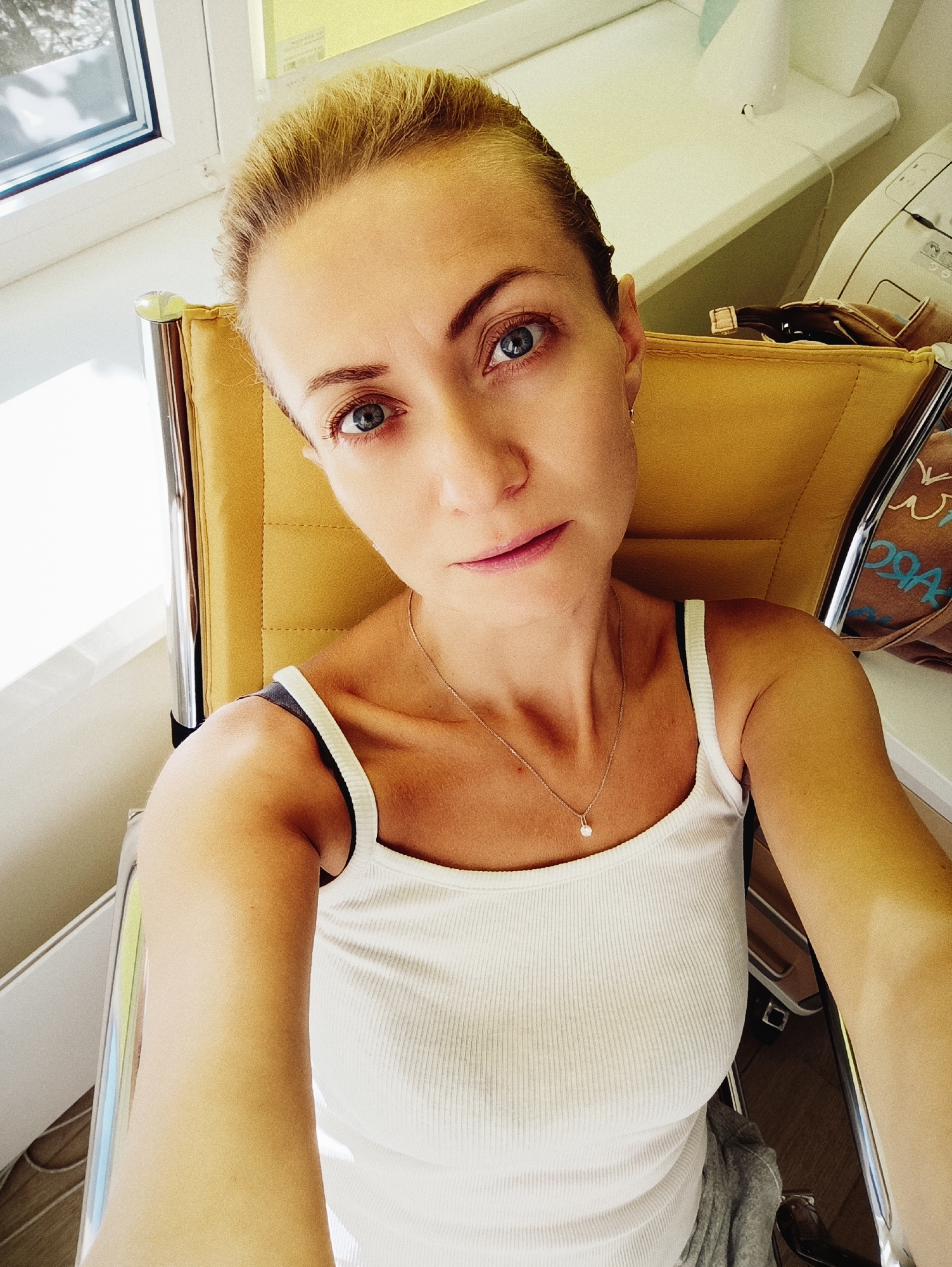 A young Caucasian woman with blonde hair pulled back is seated in a yellow chair. She is wearing a white ribbed tank top and a delicate silver necklace with a small pendant. The woman is looking directly at the camera with blue eyes. She is positioned in what appears to be an office or waiting area, with a window visible to her left. In the background, there is a white desk or counter with some equipment, and a brown bag with text on it. The overall lighting suggests it is daytime. The location is not identifiable as Moscow, Russia.