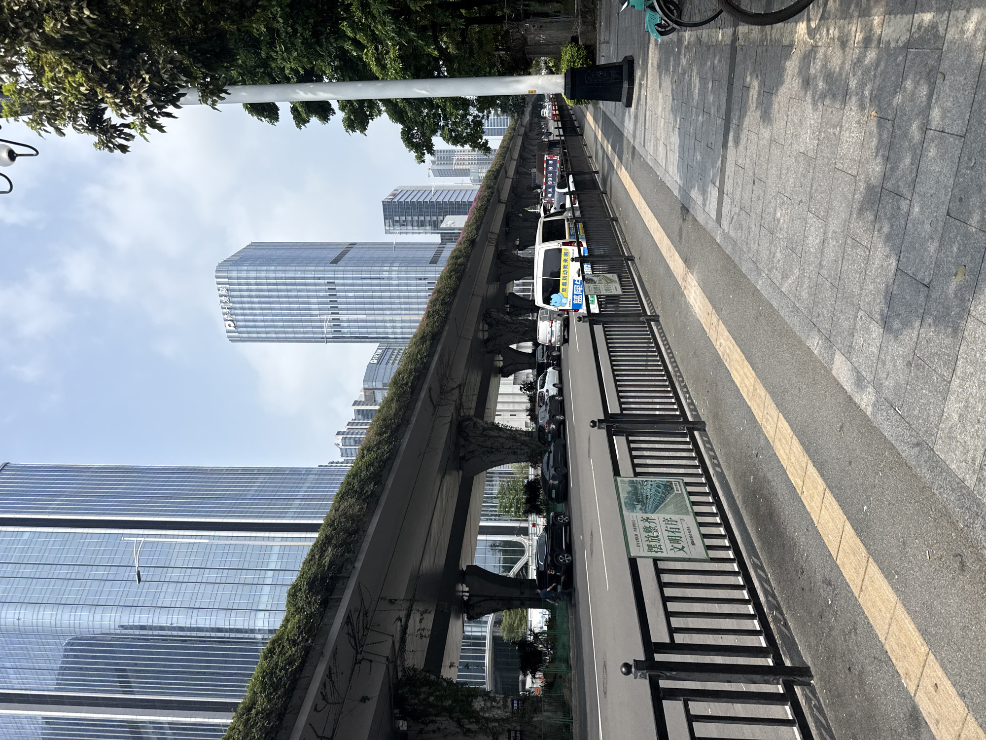 This image depicts a street scene in Huangpu District, China, likely under an elevated highway. Towering glass-fronted skyscrapers dominate the background. A long, elevated structure, covered in greenery and supported by large, vine-covered pillars, arches over the road. The road itself has multiple lanes of traffic, with cars, vans, and a police vehicle visible. A black metal fence separates the road from a sidewalk paved with grey tiles. A yellow tactile paving strip runs along the edge of the sidewalk. Several bicycles are parked on the sidewalk, and trees line the right side of the image, casting shadows on the ground. A sign with Chinese characters is attached to the fence in the foreground. The sky is clear and blue.