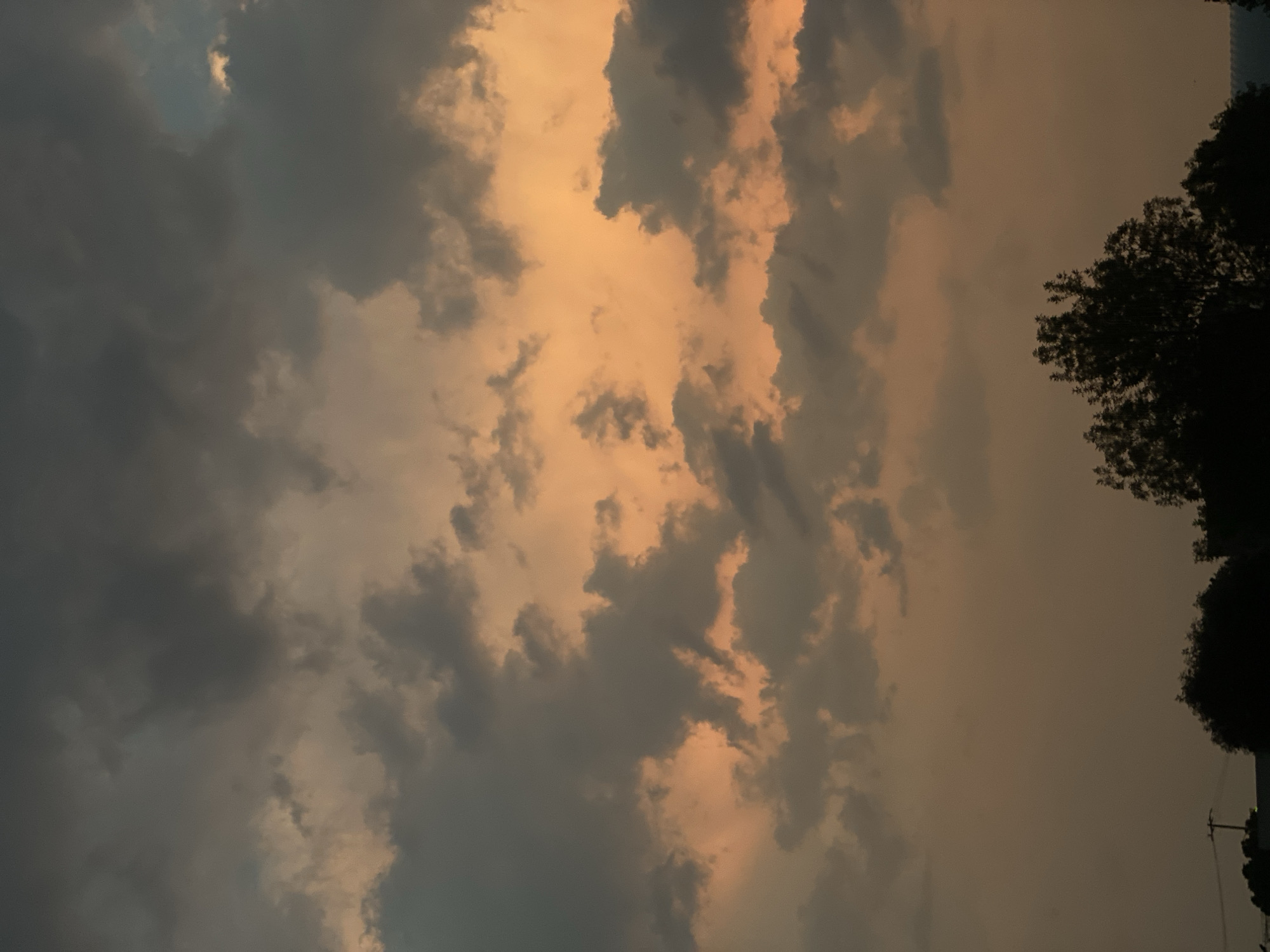 The image depicts a cloudy sky with a gradient of colors, ranging from dark gray to orange hues, likely during a sunset or sunrise. The sky is filled with textured clouds. On the right side of the frame, silhouettes of trees and utility poles are visible against the sky. The location is Unknown, Unknown.