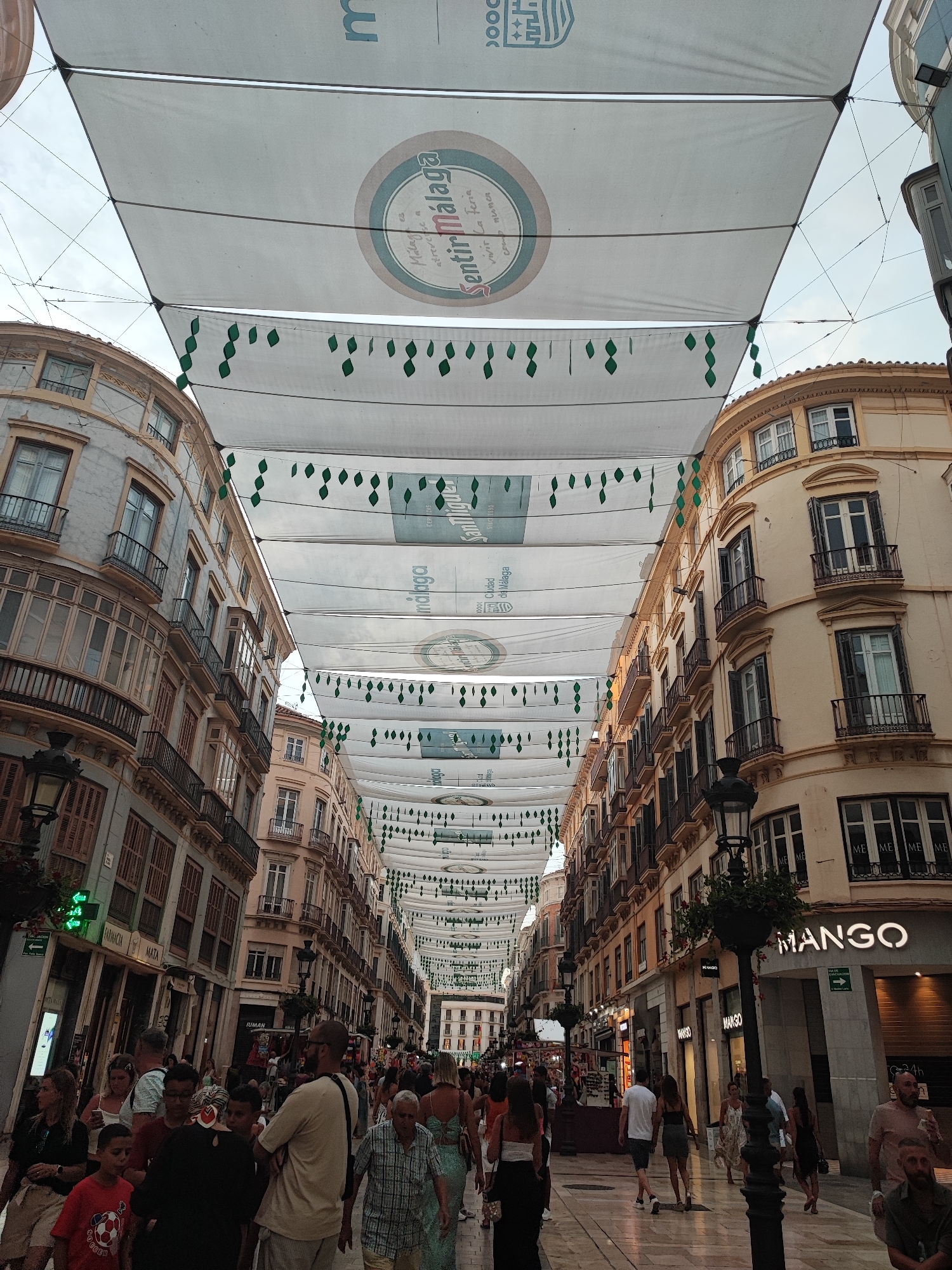 This image captures a bustling street scene in Malaga, Spain, likely during a festival or special event, given the overhead decorations. The focal point is a long pedestrian street lined with multi-story buildings on either side. These buildings feature traditional Spanish architecture with balconies, shutters, and ornate facades in shades of light blue, beige, and pale yellow.
Draped across the street at varying heights are numerous large, white fabric banners. Some of these banners display circular logos and text, including "Sentir Málaga" and "Ciudad de Málaga," indicating the city's name and a local sentiment or event. Hanging from some of the banners are green, diamond-shaped decorative elements.
The street itself is paved with light-colored tiles and appears crowded with people. Individuals are walking in various directions, some in small groups, others alone. They are dressed in casual summer attire, suggesting warm weather. Among the crowd, visible details include a person wearing a red t-shirt with a soccer ball logo, a man in a light-colored t-shirt and glasses, and several women in dresses and tops.
On the ground level of the buildings, numerous shops and businesses are visible, with illuminated signs and storefronts. Notably, a prominent "MANGO" sign is present on one of the buildings. Streetlights and lampposts are interspersed along the sidewalk. The overall atmosphere is one of lively activity and urban commerce. The perspective is looking down the street, with the sky partially visible through the gaps in the overhead banners.