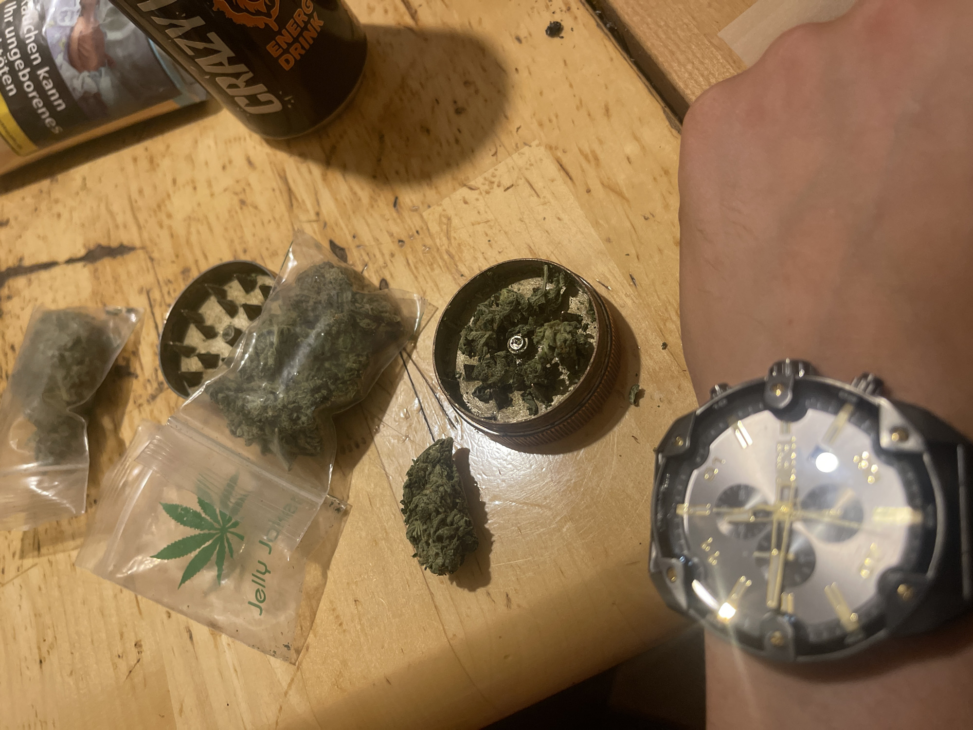 On a wooden surface are various items. There's a grinder, both empty and filled with what appears to be cannabis. Two plastic bags contain more of the same substance, with one bag labeled "Jelly Joker" and displaying a cannabis leaf. Also present is an energy drink. A wrist wearing a large watch is visible. The location is unknown.