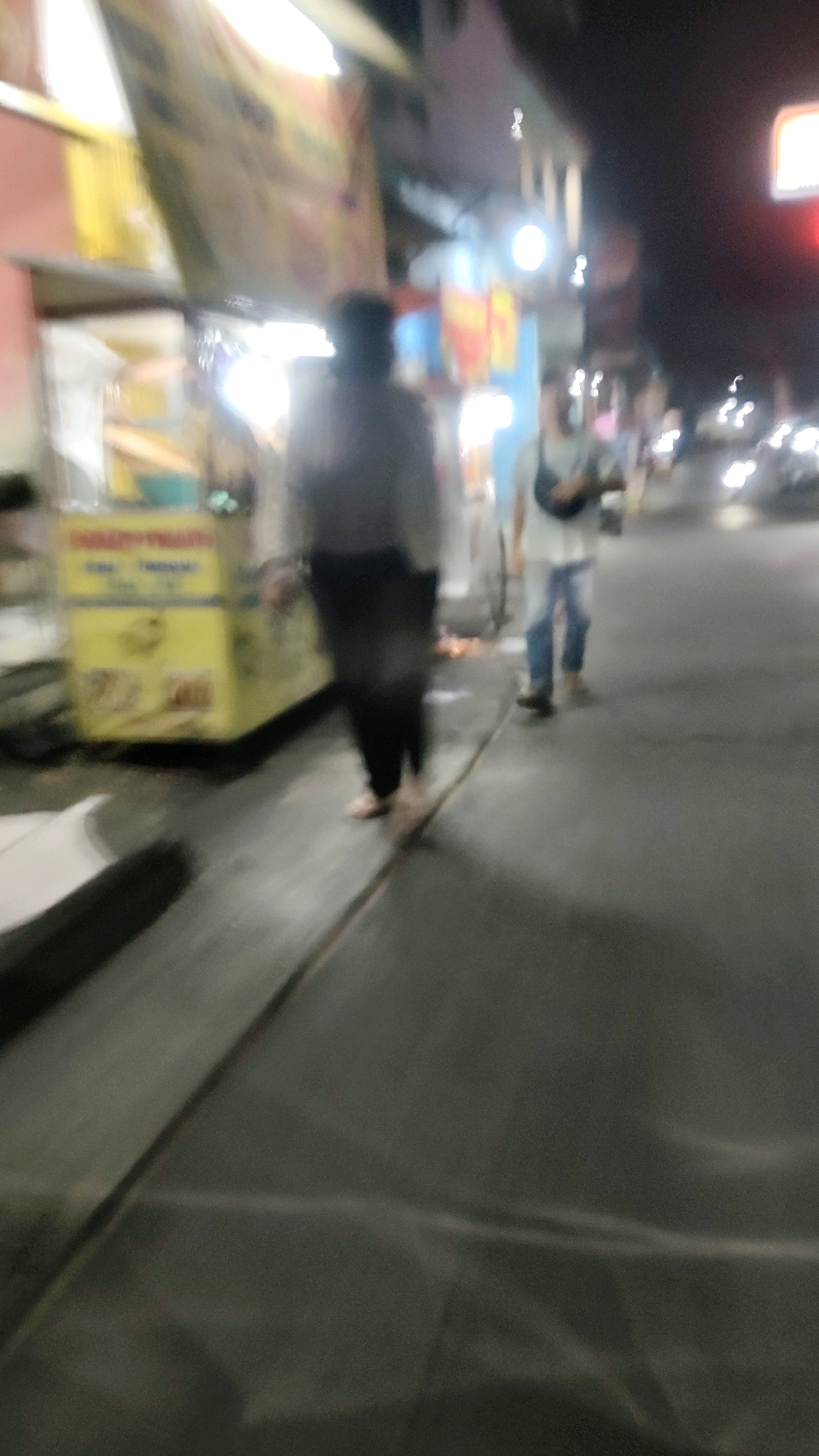 A blurred, night-time street scene in Mekarmukti, Indonesia, shows two individuals walking on a sidewalk beside a food cart. The person on the left, closer to the cart, is wearing dark pants and a light-colored, possibly gray, top. The person on the right is wearing blue jeans and a light-colored t-shirt, with a dark bag slung across their body. They are both walking away from the camera. The food cart, which is yellow with signage, is illuminated by overhead lights. The street is paved, and the lighting suggests it is a commercial area. Other blurred figures and vehicle lights are visible in the background, indicating ongoing street activity.