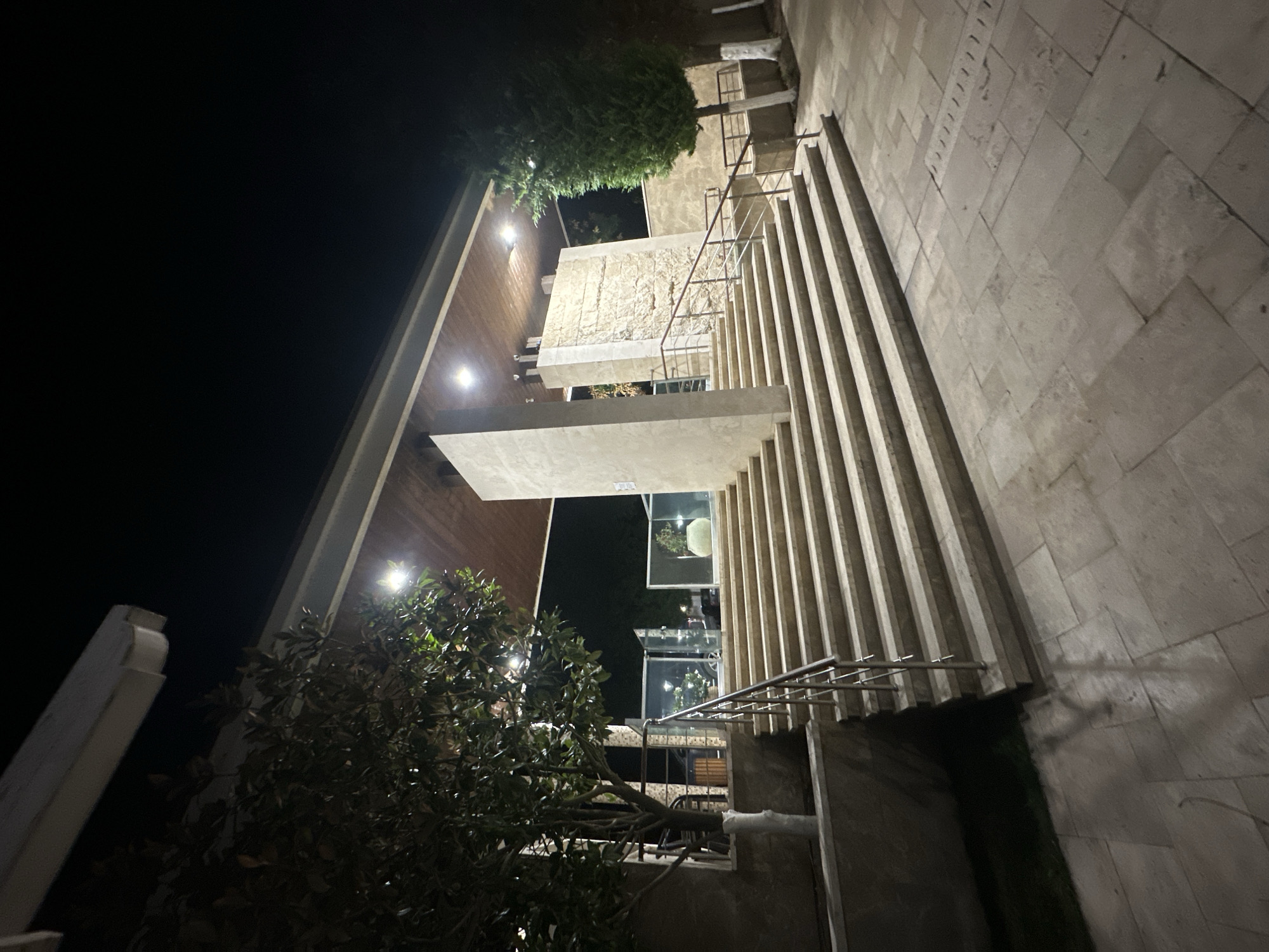 A nighttime, eye-level, full shot shows a wide stone staircase leading up to a building entrance at the city of Sozopol, Bulgaria. The staircase is flanked by metal railings and a large tree on the left. The entrance area is covered by a wooden roof illuminated by bright overhead lights. Behind the staircase, there is a textured stone wall with what appears to be ancient carvings. To the right of the stairs, another tree stands tall. The surrounding ground is paved with large stone tiles, and shadows are cast on the ground from the lighting. The sky above is dark, indicating nighttime. There are no people clearly visible in the image, but the setting suggests a public or commercial space.