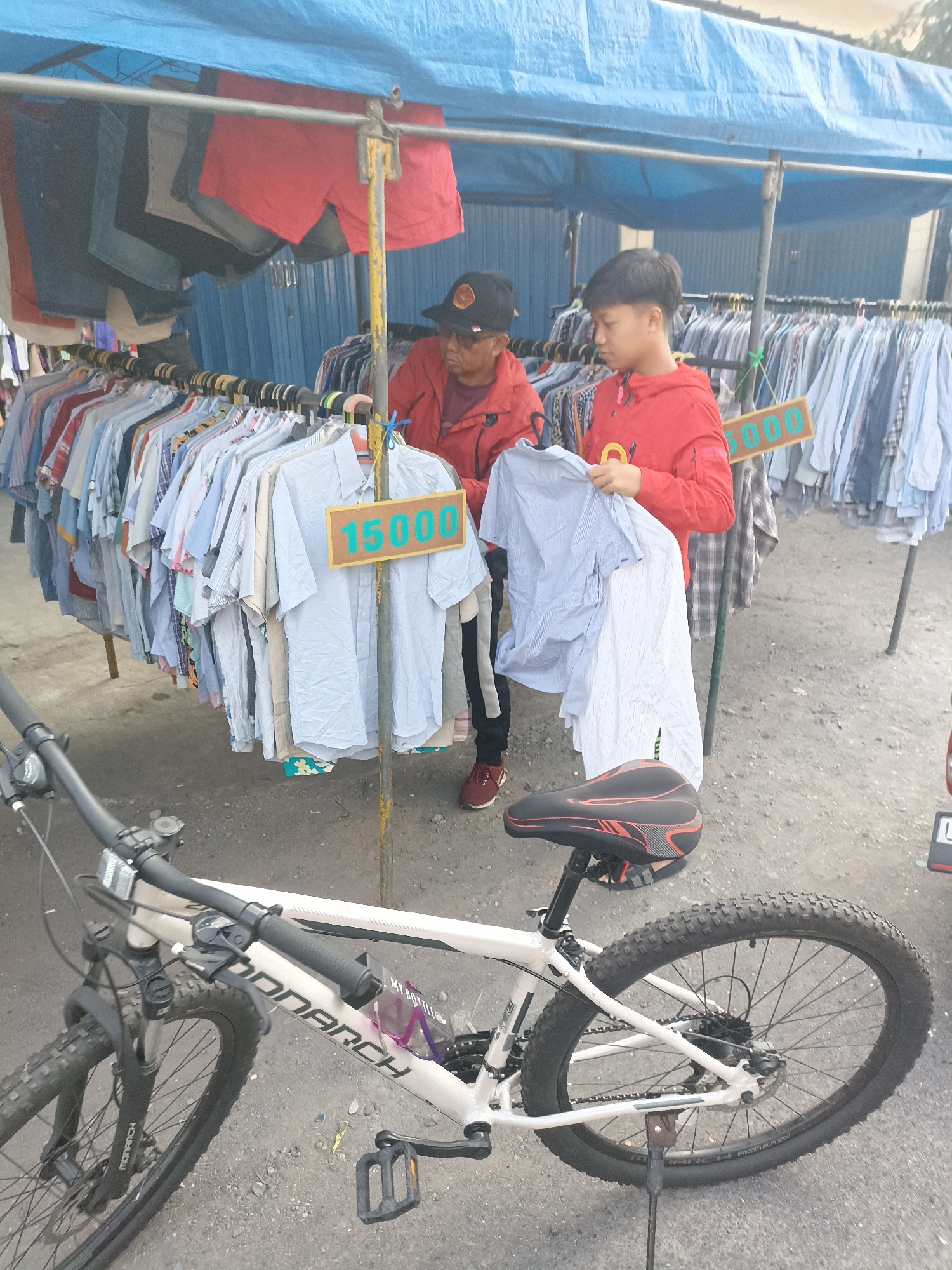A man and a boy are at an outdoor clothing stall in Surabaya, Indonesia. The man, wearing a red jacket and a dark baseball cap, looks at clothing on a rack. The boy, also in a red jacket, holds up a blue and white striped shirt. Behind them, racks of shirts are displayed under a blue tarp. A sign with "15000" is visible on a pole. In the foreground, a white bicycle with black tires is parked. The ground is rough and dusty.