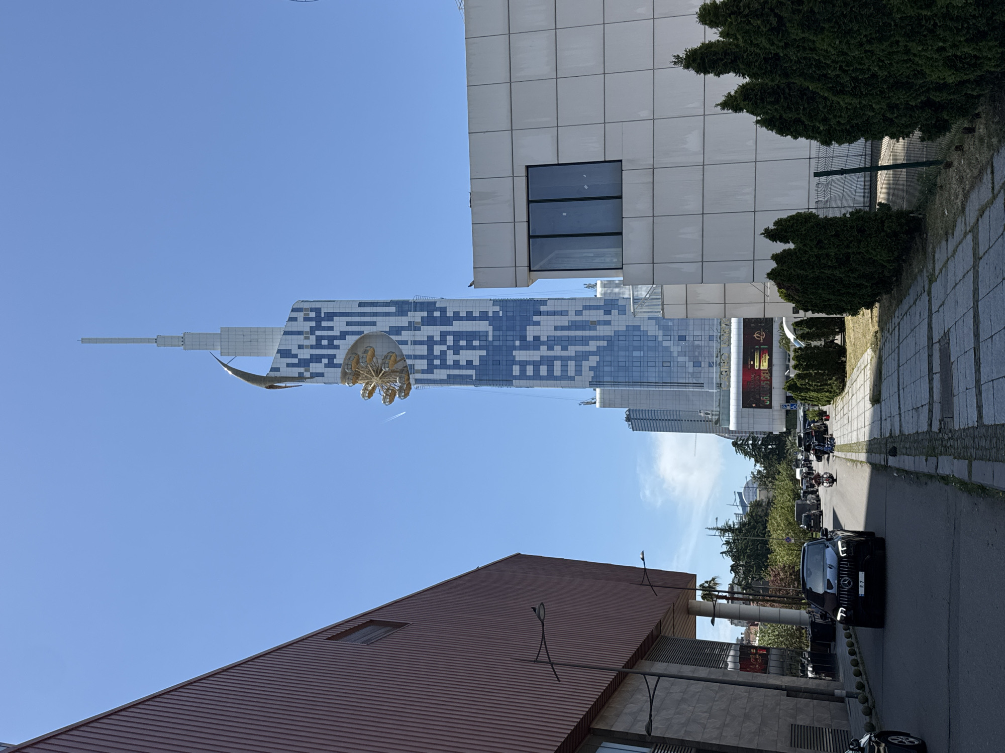 The image captures a street scene in Batumi, Georgia, featuring several buildings and vehicles. In the background rises the distinctive silhouette of the Batumi Tower, an architectural landmark known for its design mimicking a DNA double helix and topped with a Ferris wheel. Adjacent to the street is a modern building with a white and grey tiled facade and a prominent window on the upper level. Trees and paved walkways line the side of the street. A black car is visible on the road, with a line of motorcycles following behind. A building with a brown roof is present in the foreground. The sky is clear and blue.