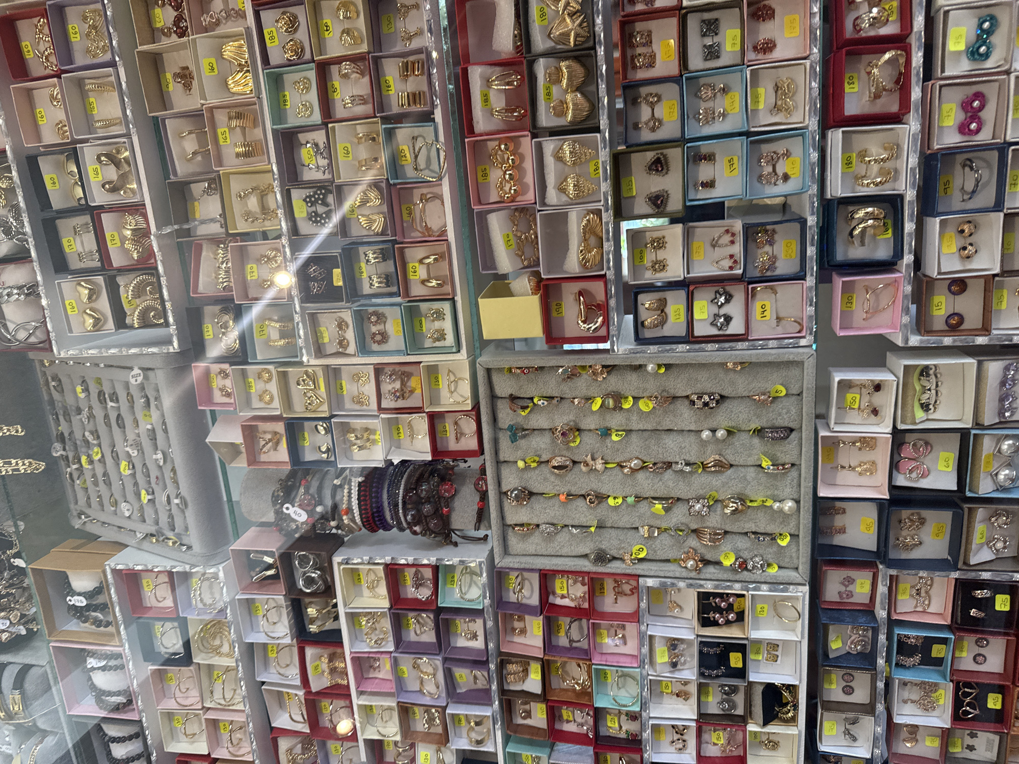 This image depicts a large display of jewelry, likely for sale. The jewelry consists of rings, earrings, necklaces and bracelets organized in small boxes, arranged closely together, covering a significant area, likely a store shelf or display case. The jewelry varies in style and color, including gold, silver, and colored stones. Prices are indicated on the jewelry and packaging. The location of this image is the city of San Cristóbal, Mexico.