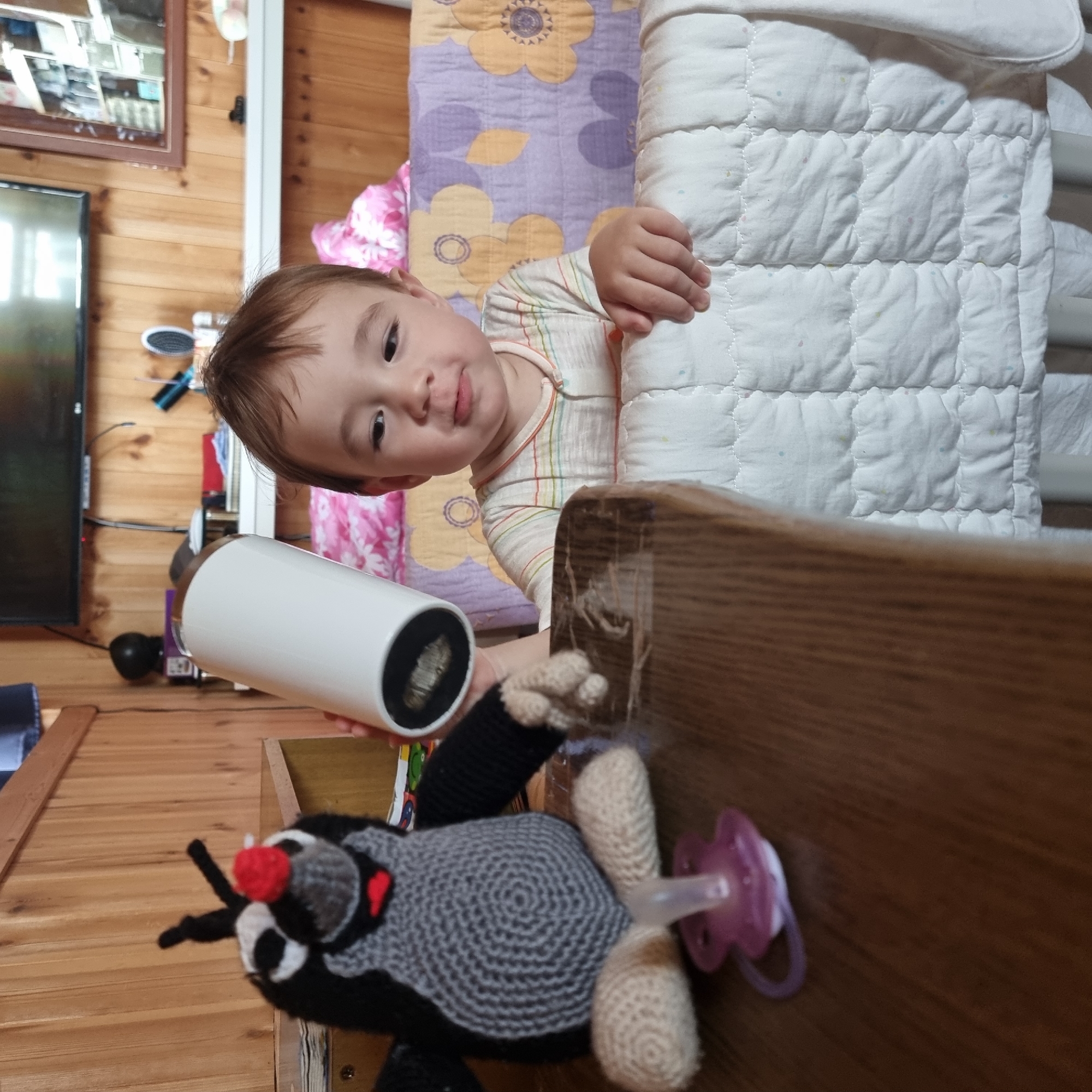An infant boy with brown hair and a striped shirt is sitting in a crib. He is looking to his left, towards a crochet toy mole and a white cylinder held by an unseen person. The toy mole is grey and black with a red nose and a pink pacifier in front of it. The crib has a white quilted mattress and colorful bedding. The background appears to be a room with wooden walls and a television. The image does not provide any information about the location of Dunnae-myeon, South Korea.