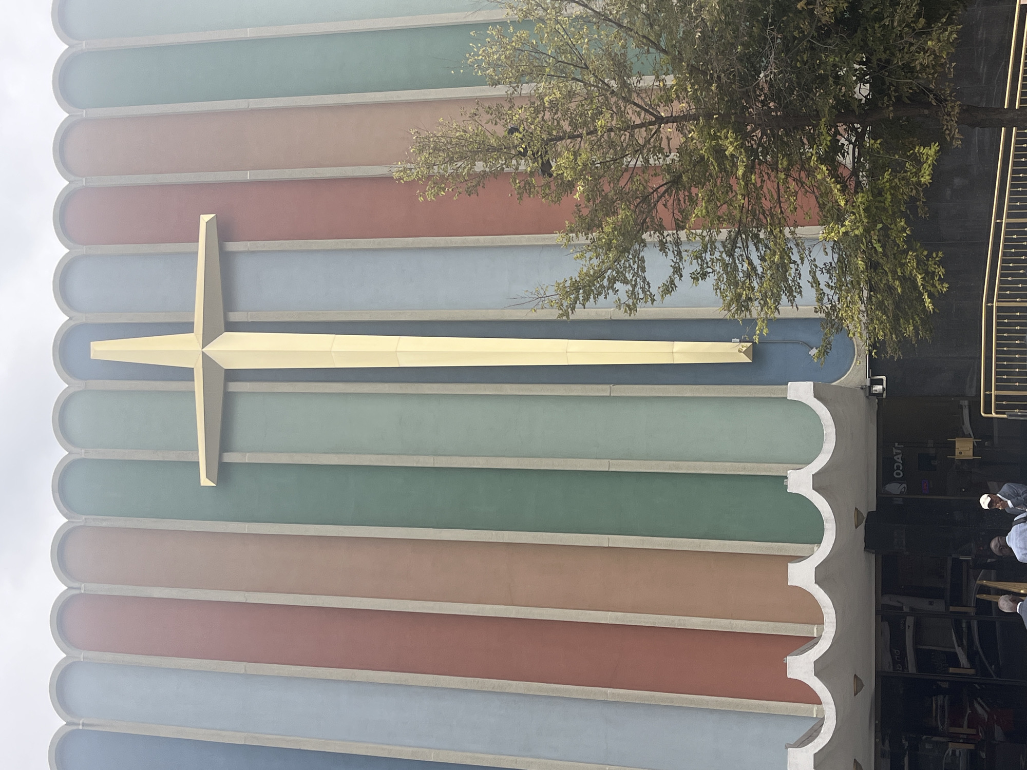 An exterior, eye-level, medium shot captures the colorful facade of the First Church of God in Christ located in the Harlem neighborhood of New York City, USA. The building features a prominent, golden, three-dimensional cross as its centerpiece, set against vertical panels of varying hues, including blue, light blue, red, peach, green, and light green. A lush green tree with dense foliage partially obscures the lower right portion of the building. At the bottom of the image, the entrance to the church is visible, with a scalloped white overhang above dark glass doors. Three men are standing near the entrance, two of them facing away from the camera and one looking towards it. A golden railing is visible in the foreground.