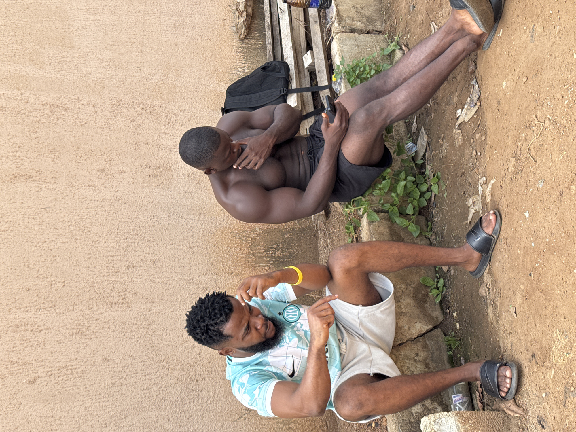 The image shows two men outdoors in what appears to be a casual, possibly gritty setting. One man, shirtless and wearing black shorts, is sitting with his legs extended, holding a smartphone. He has a muscular build and is looking down at his phone, with one hand resting on his chin. Behind him, a black backpack rests on a wooden structure. The other man is sitting with his legs bent and is wearing a light blue and white patterned t-shirt and light-colored shorts. He has a beard and dreadlocks, and is looking towards his right, gesturing with both hands. He is wearing a yellow wristband on his left arm. Both men are wearing sandals.
The ground is a mix of dirt and cracked concrete, with some greenery and scattered debris. The wall in the background is a textured, sandy color. The lighting suggests it is daytime, and the overall atmosphere is relaxed. There is no visible text in the image. The location context is Akwanga, Nigeria.