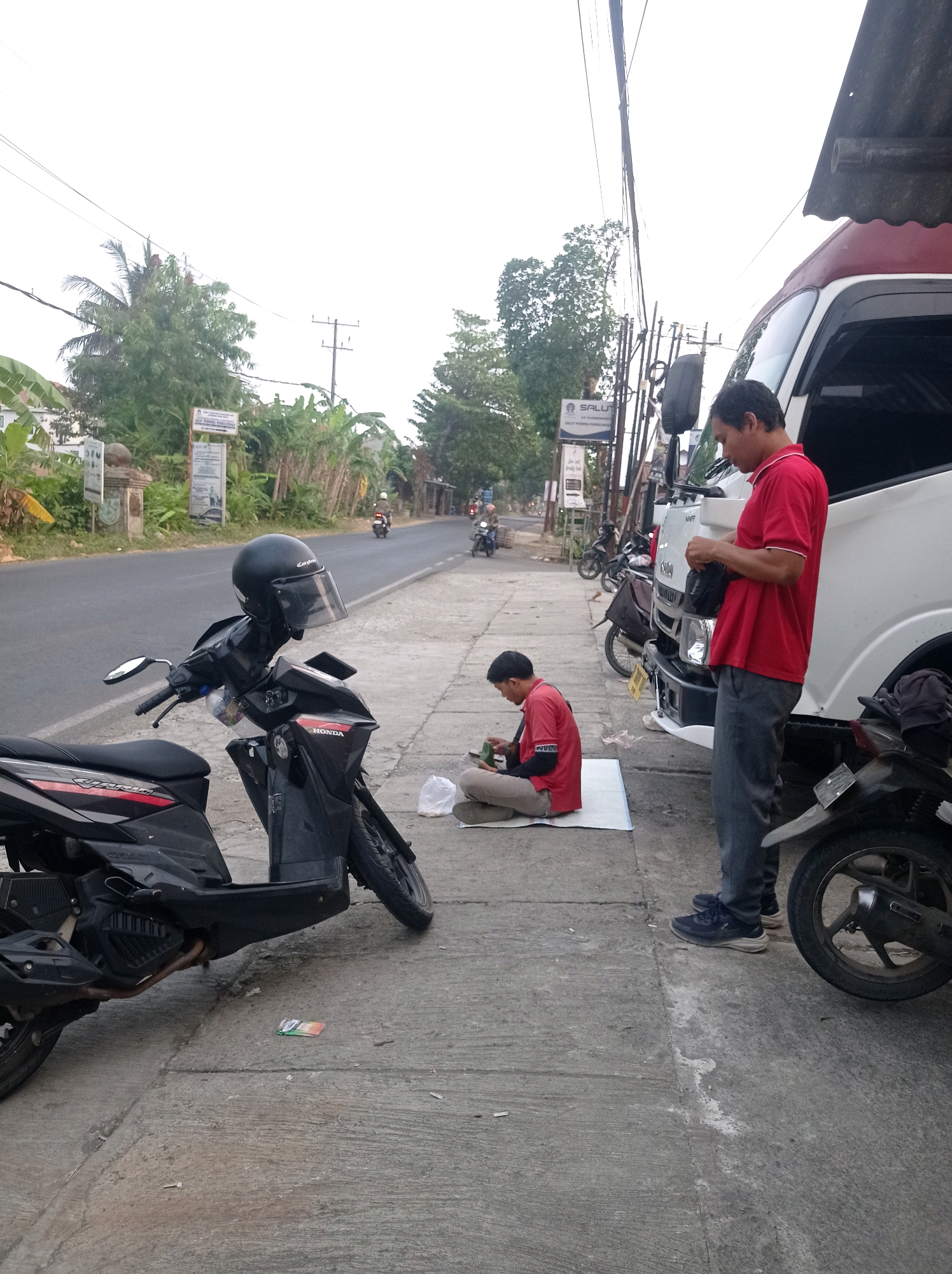 In Jetis, Indonesia, two men are present on a sidewalk next to a road. A black scooter with a black helmet on its handlebars is parked on the sidewalk in the foreground. To the right, a white truck is parked. One man, wearing a red shirt and grey pants, stands beside the truck, holding a dark bag and looking down. Another man, also in a red shirt and grey pants, is seated on a white mat on the sidewalk, looking at a phone. The road behind them shows two-wheeled vehicles and some buildings. The environment is urban, with overhead wires and vegetation visible.