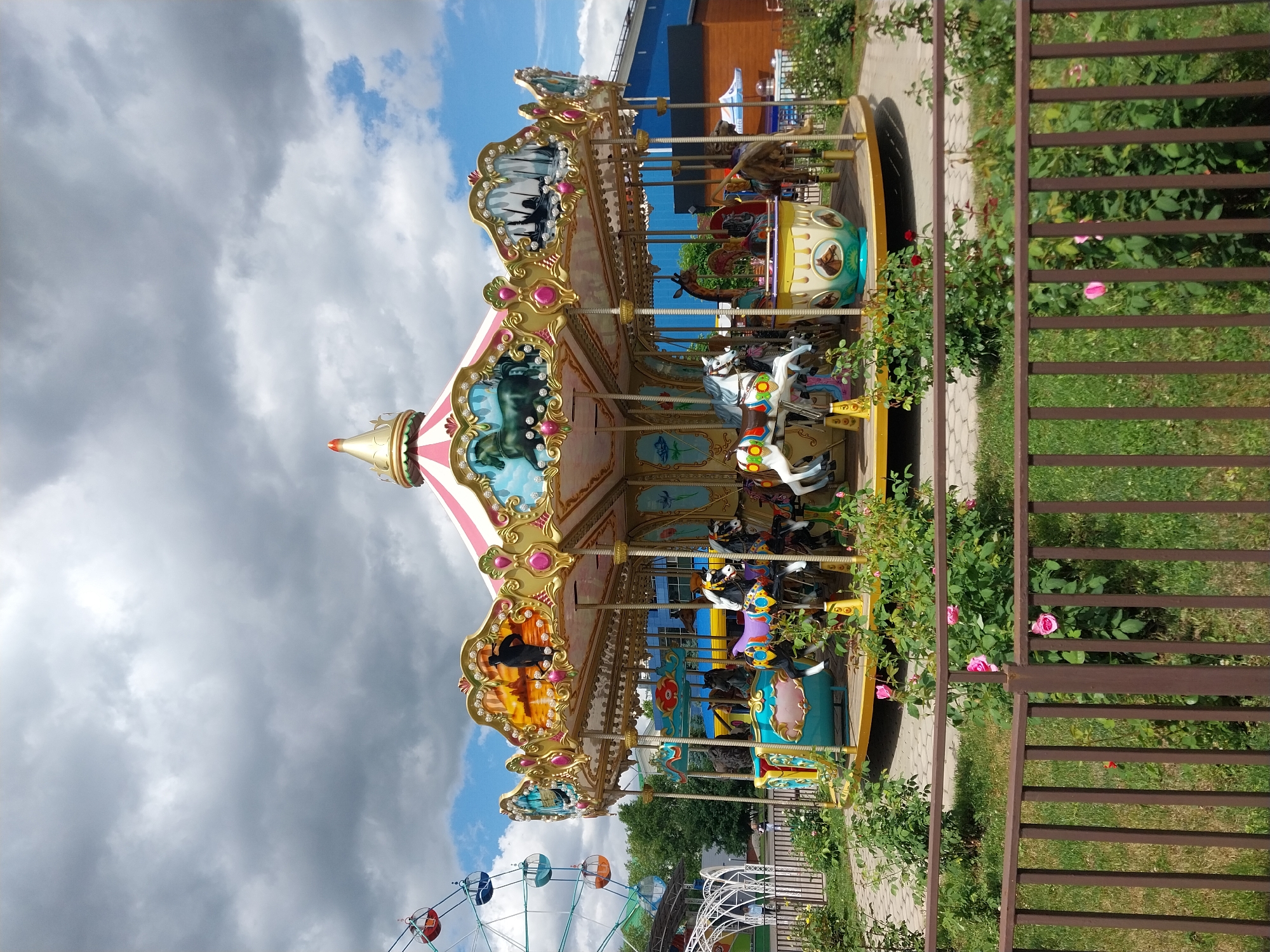 The image shows a carousel in a park, potentially in Orenburg, Russia based on context clues. The carousel is multi-tiered, brightly colored with decorative elements including animals and landscape scenes on its panels. The top of the carousel is striped in pink and white, topped with a gold finial. The structure itself has horses and other animals. A Ferris wheel is visible in the background. A brown metal fence borders the right side of the image. Green vegetation with pink flowers is adjacent to the fence and the carousel. The sky above is partly cloudy with patches of blue. The photograph appears to be taken from a slightly low angle.