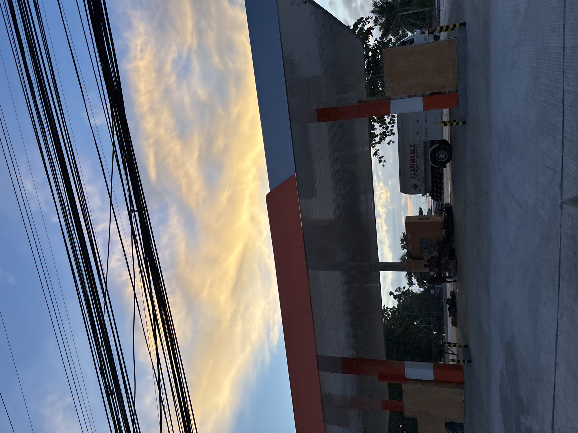 The image depicts a gas station under construction at dusk in Davao City, Philippines. A large, empty gas station canopy dominates the foreground, with orange and blue accents on its roof. Power lines are strung overhead, crisscrossing the sky, which is painted with soft hues of blue and orange from the setting sun.
In the middle ground, several construction workers are visible near a small orange structure, possibly a kiosk or an office. They appear to be engaged in activity, though their specific tasks are unclear. A tanker truck, labeled "FLAMMABLE" in bold red letters, is parked to the right of the workers, its trailer white with black lettering. Construction materials, including wooden boards, are stacked around the site, suggesting ongoing work. Palm trees and other foliage are visible in the background, indicative of the tropical climate. The ground is paved with concrete, with some areas covered in what appears to be a light grey asphalt. The overall atmosphere is one of a developing urban landscape at the end of the day.