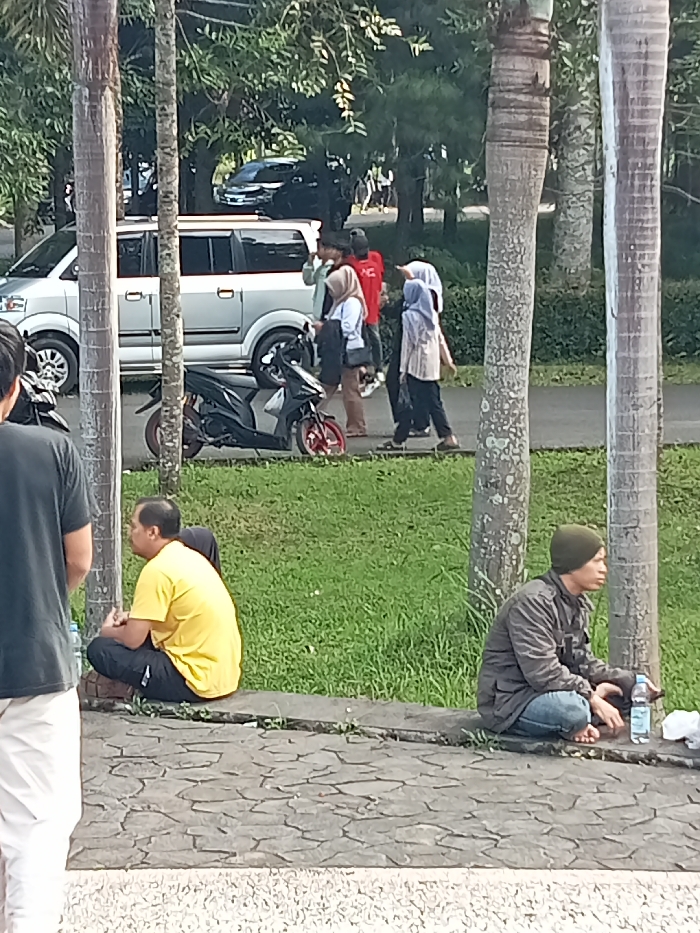 This outdoor scene, likely captured in Singaparna, Indonesia, depicts everyday life with several people and vehicles amidst green foliage under bright daylight.
In the foreground, on the left, the back of a person wearing a dark t-shirt and white pants is partially visible, with a water bottle beside them. To their right, a man in a yellow t-shirt and dark pants squats on a low, paved curb. Further right, another man in a green beanie, grey jacket, and blue jeans squats barefoot on the ground next to a palm tree, with a water bottle and a white wrapper nearby.
The midground features a silver minivan, resembling a Suzuki APV, parked or moving slowly. Next to it is a black scooter with red accents on its wheels. A group of five individuals walks along a paved path to the right of the vehicles. This group includes people dressed casually; some women are wearing hijabs (headscarves), and one person wears a red t-shirt. They appear to be walking in a relaxed manner.
The background is dominated by lush green grass, numerous tall palm trees, and dense greenery, suggesting a park-like or verdant roadside environment. Other vehicles are partially visible further back among the trees. The lighting indicates a clear day, possibly morning or late afternoon, with a generally calm and casual atmosphere. No distinct or legible text is visible on any objects or clothing.