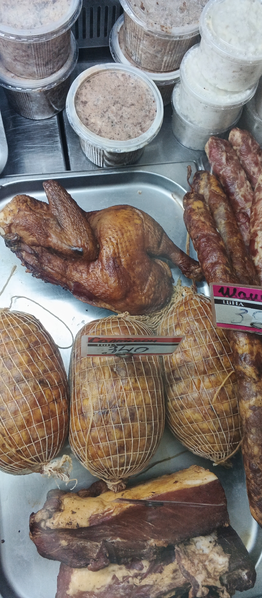 This image depicts a refrigerated display case containing various food items for sale at a market in Ploske, Ukraine. The display includes several clear plastic containers filled with what appears to be processed meat or pâté, as well as some white, creamy products. In the foreground, a whole roasted chicken is presented on a metal tray. Next to the chicken are several cylindrical food items wrapped in netting, some with price tags. A portion of raw, cured meat is visible at the bottom of the image. The overall setting suggests a food stall or a section of a larger grocery store specializing in meat products.