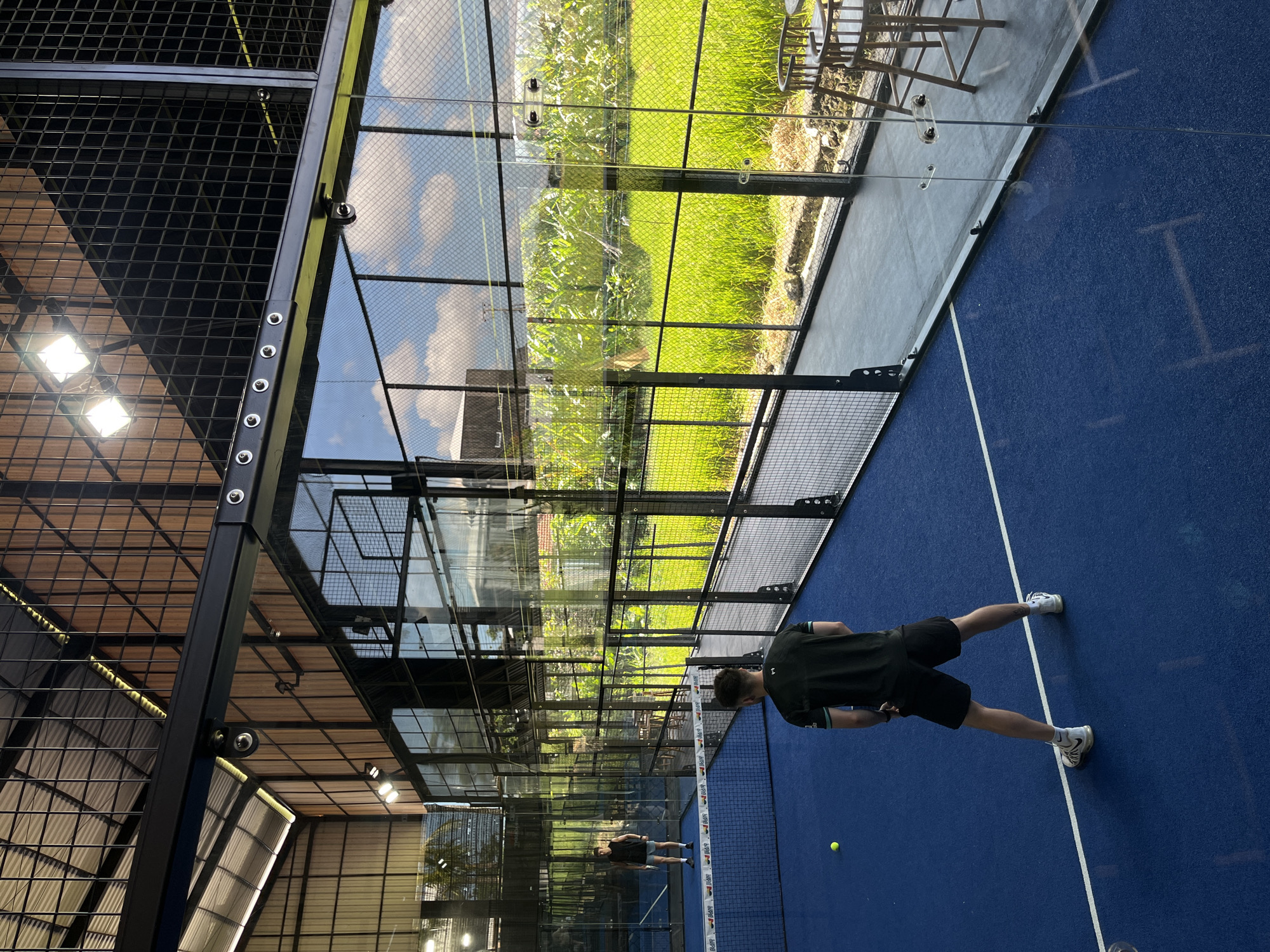 The image depicts a padel court in North Kuta, Indonesia, where two men are engaged in a game during daytime. The court has a vibrant blue playing surface marked with white lines, and a yellow padel ball is visible on the court in the foreground.
The player closer to the viewer is positioned on the right side of the court, seen from behind, wearing a dark t-shirt and shorts with white athletic shoes. He stands in a ready stance, facing the net. Another player is visible further back on the opposite side of the net, also in dark attire, ready for action.
The court is enclosed by a structure comprising metal mesh walls and clear glass panels. The net separating the two halves of the court features the brand name "BARRR" repeatedly in white lettering.
The glass walls prominently reflect a bright outdoor scene, showing lush green fields or vegetation, likely rice paddies given the location, under a sky with scattered clouds. These reflections dominate the left side of the image. Concurrently, reflections of indoor lighting fixtures and the facility's ceiling structure are also discernible on the glass and mesh, particularly towards the right and top sections of the enclosure. Beyond the court's structure, some outdoor seating, including chairs and tables, is visible amidst the greenery. The setting combines aspects of an enclosed sports facility with an open view of the surrounding natural landscape.