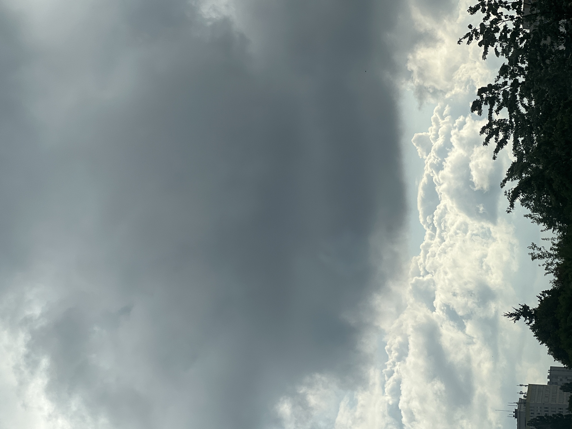The image depicts a cloudy sky with dramatic, dark grey storm clouds dominating the upper portion. Below these, a layer of brighter, puffy white clouds is visible, with patches of lighter sky peeking through. The bottom third of the image shows the silhouettes of trees and buildings against the sky. On the left, a multi-story building with an antenna structure on top is partially visible. The overall scene suggests impending rain or a recent storm. The context of Warsaw, Poland, is not discernible from the visual elements in the image.
