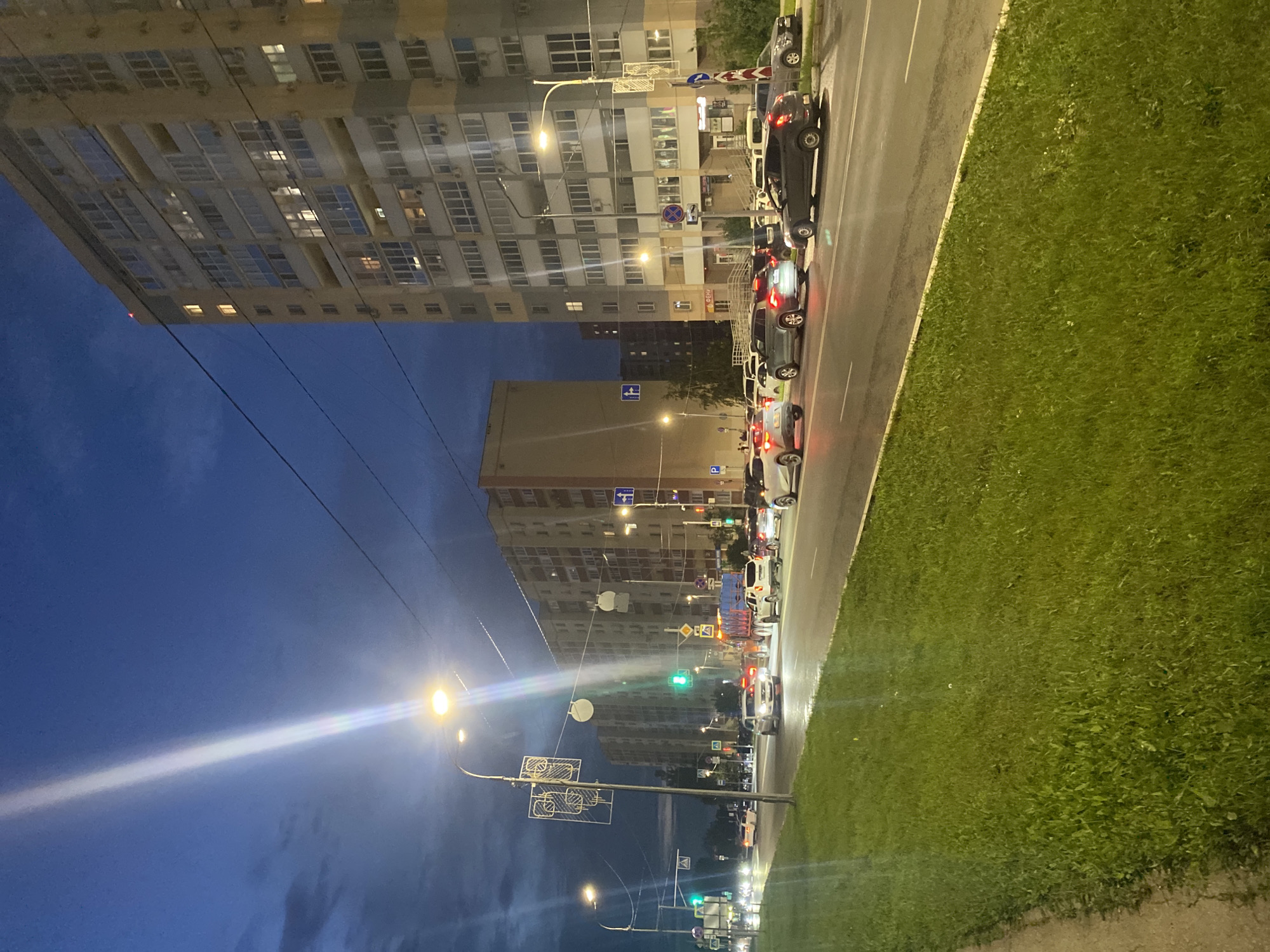 The media file depicts a street scene at dusk in Novokuybyshevsk, Russia. Tall, multi-story apartment buildings with numerous illuminated windows line the left side of the frame. A row of cars is parked or driving along the right side of the asphalt road, with their headlights and taillights visible. A grassy area borders the road on the right. Streetlights are illuminated, casting a warm glow on the scene, and a bright streetlight on the left creates a strong lens flare. The sky is a deep blue, transitioning from day to night, with some clouds. Traffic lights are visible, with one showing a green light. Various traffic signs are also present. The overall atmosphere suggests a typical urban evening with activity on the street. There are no people clearly visible.