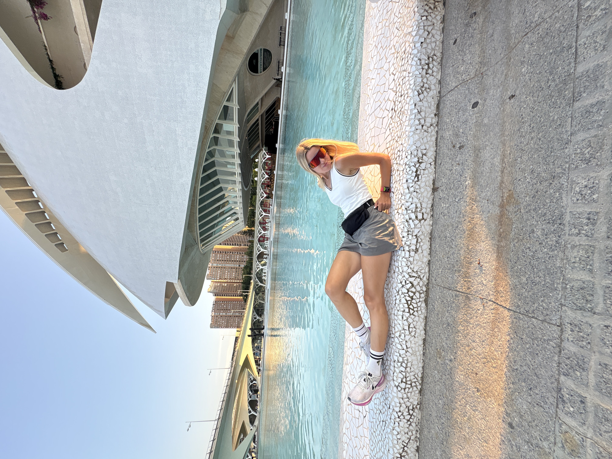 A blonde woman with red-tinted sunglasses is lying on a white tiled ledge next to a bright blue pool of water. She is wearing a white tank top, grey shorts, white socks with black stripes, and white athletic shoes with pink accents. Her body is angled towards the camera, with her legs extended. Behind her, a futuristic white building with curved architectural elements rises against a clear blue sky. In the background, a modern bridge with multiple arches spans across the scene, and several tall residential buildings are visible. The setting appears to be the City of Arts and Sciences in Valencia, Spain, a complex of futuristic buildings designed by Santiago Calatrava. The sun is low in the sky, casting long shadows and a warm glow on the ground in the foreground.