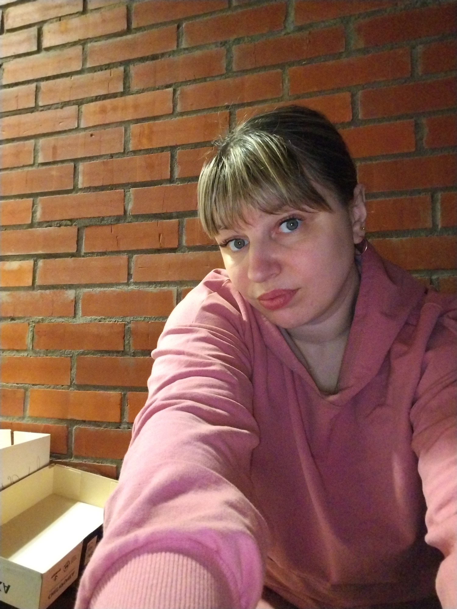 A woman with light brown hair and blonde highlights, styled with bangs across her forehead, is featured in this close-up indoor portrait, likely a selfie. She has blue eyes and is wearing subtle makeup, including eyeliner and lipstick, which gives her lips a slight pout. Her expression is neutral or mildly pensive. She is dressed in a casual pink hooded sweatshirt with a cowl neck. Small hoop earrings are visible on her ears.
The background consists of a reddish-orange brick wall with grey mortar, suggesting an interior space with exposed brickwork. The lighting appears warm and artificial. In the lower left foreground, two open, light-colored cardboard boxes are partially visible. Text in Cyrillic is visible on the boxes; "ЧЕЛАКОМ" (CHELAKOM) is discernible on the larger box, and "АЗ" (AZ) is on the side of the smaller, upper box. The angle of the shot, with her arm extended towards the camera, indicates she is holding the device herself. The scene depicts a casual, possibly domestic setting in Шебунино, Russia.