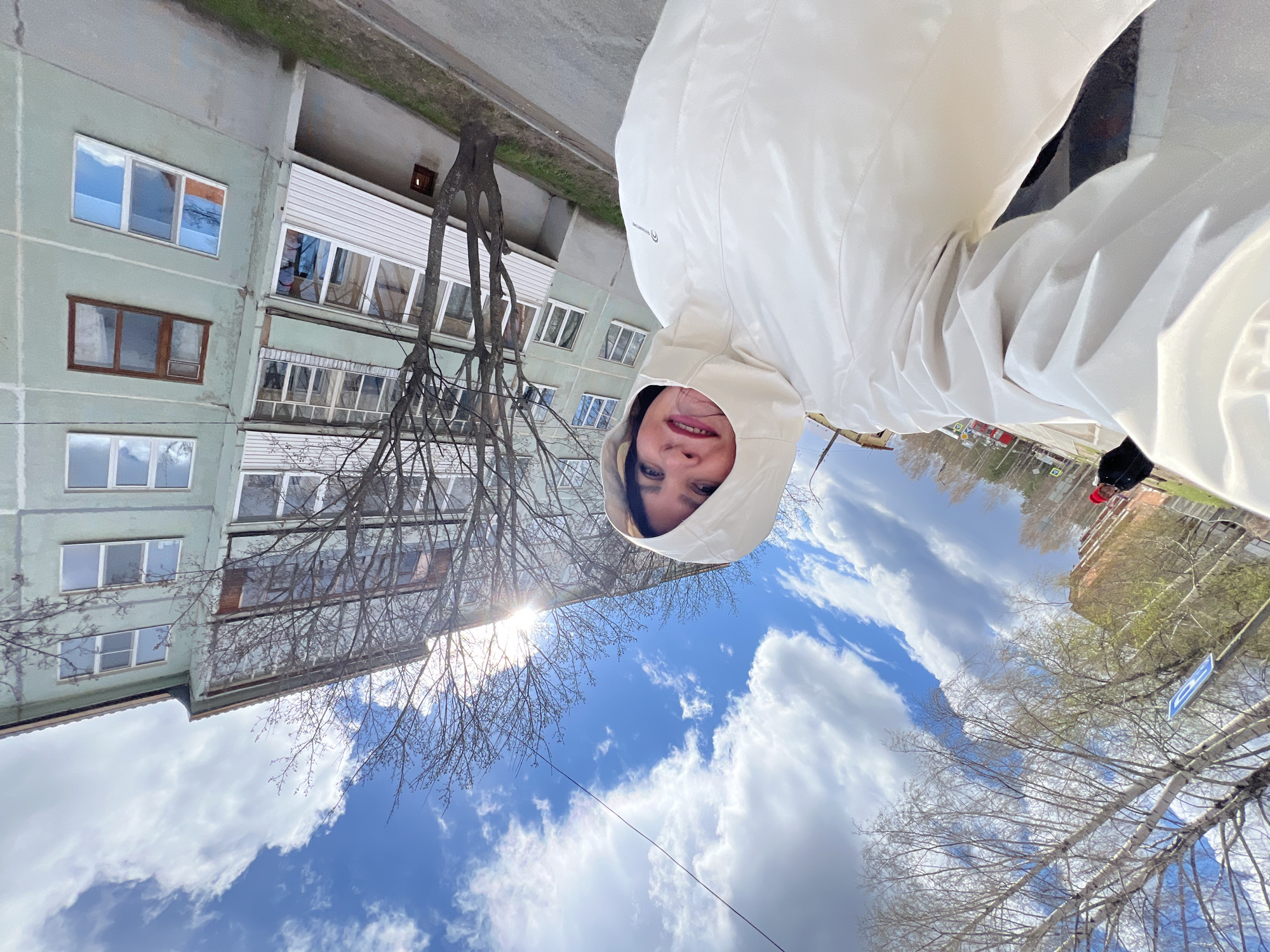 The image is oriented vertically and rotated 90 degrees counter-clockwise, presenting an unusual perspective. A person, appearing to take a selfie, is positioned on the right side of the frame. They are wearing a white, slightly shiny hooded jacket, with their face framed by the hood. Dark eyes and a slight smile are visible. On the jacket's left chest area, there is a faint black logo, possibly including the word "ADVENTURE."
To the left and behind the person stands a multi-story residential building with light green-grey walls. Numerous windows, some reflecting the bright sky and clouds, are visible. One window on the upper floors has a brown frame, while most others are white-framed. Balconies or loggias, some enclosed with white vertical siding, extend from the building.
Bare-branched deciduous trees, indicative of early spring or late autumn, are prominent in the midground and foreground, partially obscuring the building. The large roots of one tree are visible near the building's base.
The lower and right portions of the frame capture a bright blue sky filled with white and grey cumulus clouds. A strong sun glare shines through the upper-left branches of a tree, suggesting bright daytime conditions. A thin power line stretches across the sky. In the far distance, beyond the trees, hints of other buildings, green foliage, and a blurry blue square sign with a white symbol are faintly discernible. The scene takes place in Noviy, Russia.