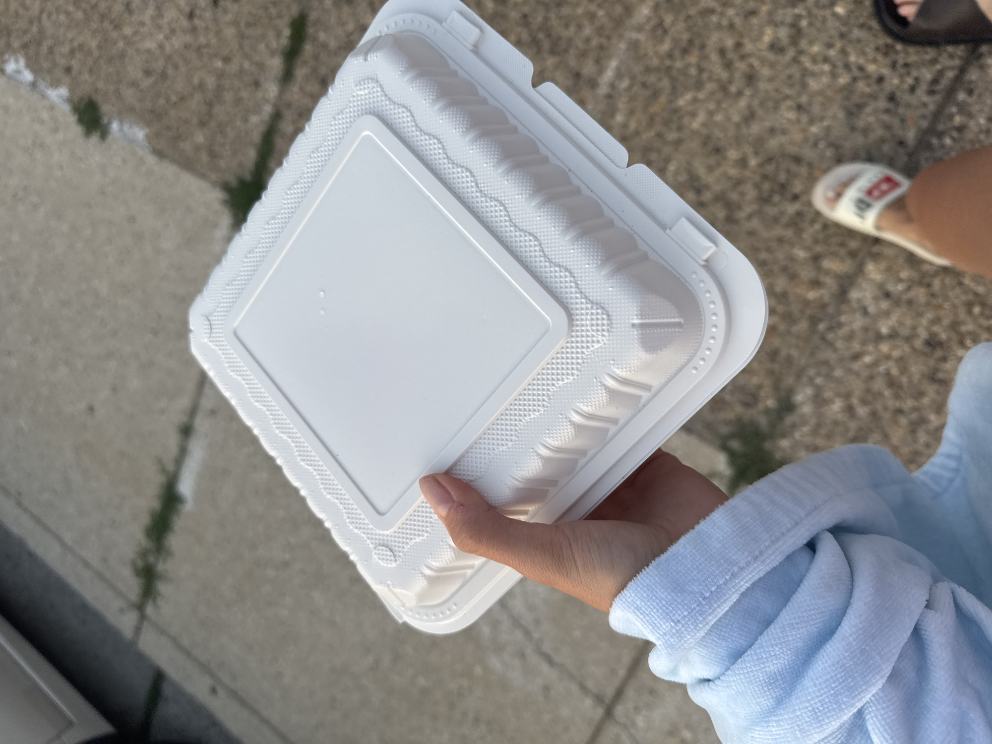 An overhead shot shows a person's hand holding a white square takeout container. The container has a ridged rim and a slightly raised, textured lid. The background is out of focus and appears to be a paved surface with some patches of grass, possibly a sidewalk or driveway. The person is wearing a light blue, terrycloth sleeve on their left arm. In the lower right corner, a blurry foot wearing a white slide sandal with a red and white logo is visible. The location is Ocean City, United States of America.