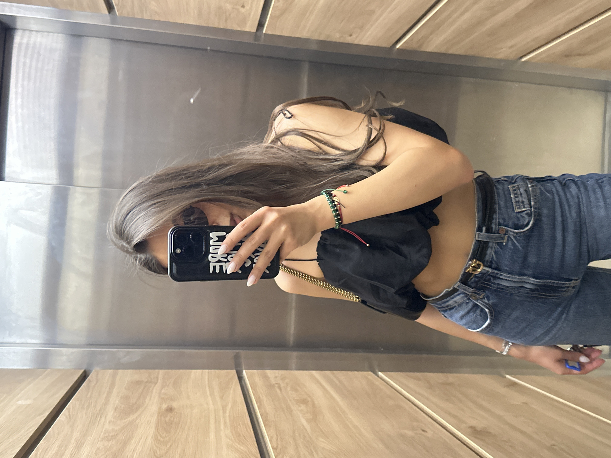 A young woman with long brown hair and sunglasses is taking a mirror selfie in an elevator. She is wearing a black crop top, blue denim jeans, and a black belt with a gold buckle. She is also wearing bracelets and has a gold chain purse slung over her shoulder. The elevator has wooden paneled walls and a metallic door. The selfie is taken at the residential building "Panorama Sofia", located in Sofia, Bulgaria.