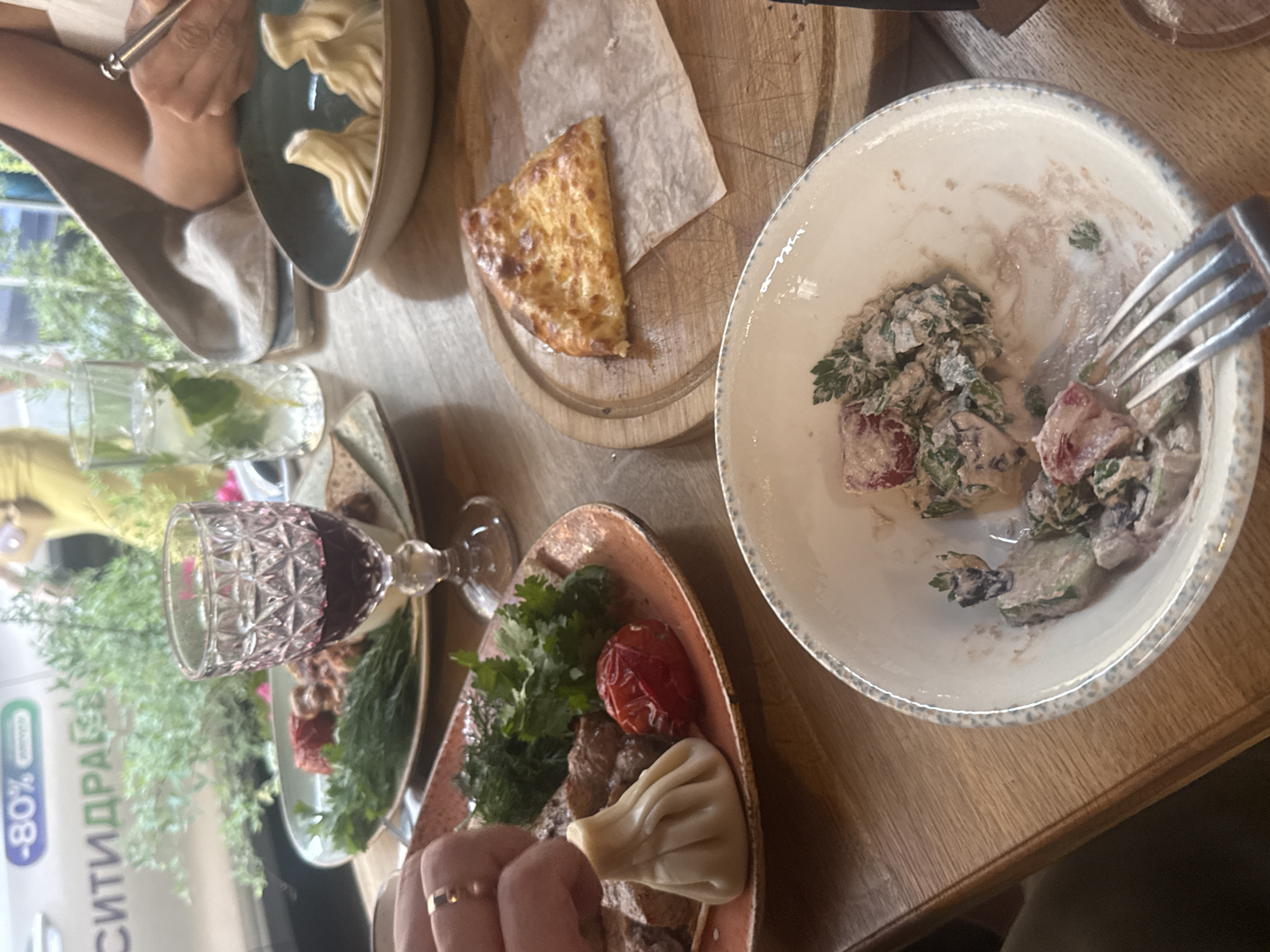 The image captures a close-up view of a wooden table laden with a Georgian meal. In the foreground, a partially eaten salad sits in a white bowl, with a fork resting on its rim. To the right, a triangular piece of khachapuri, a Georgian cheese-filled bread, is placed on a wooden board. Further back, a plate holds several khinkali, a type of Georgian dumpling, alongside grilled meat and tomatoes. A glass of red wine and a tall glass with a drink and mint are also visible on the table. In the background, a window shows a glimpse of greenery and a sign that appears to be in Russian, with the text "-80% СИТИДРАЙВ". The presence of the "-80% СИТИДРАЙВ" sign strongly suggests this scene is located in Moscow, Russia, as "Citydrive" is a carsharing service operating in Moscow.