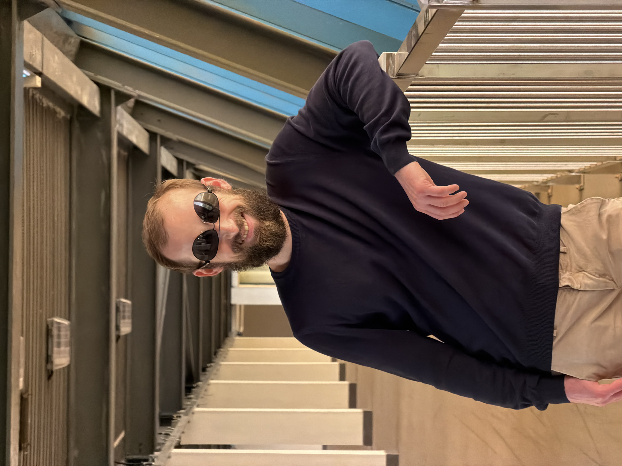 A bearded man wearing sunglasses and a navy blue sweater stands on a staircase. The image appears to be rotated. The architectural details suggest a modern interior with metal beams, lighting fixtures, and a staircase with white steps. The setting seems to be within the city of Moscow, Russia, based on the modern architectural style and possible use of Russian materials.