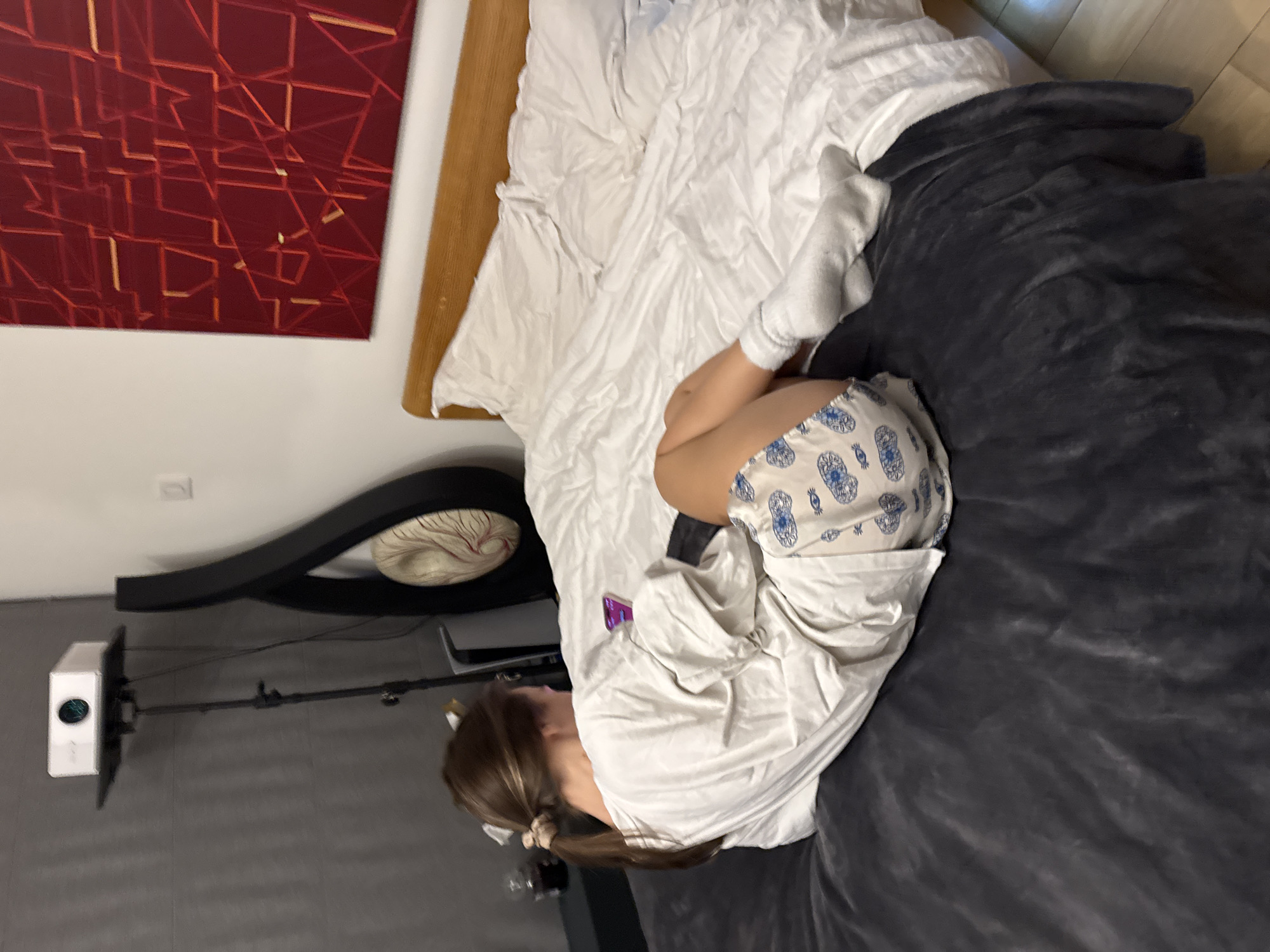 A young person is curled up on a bed, wearing socks and shorts with a pattern. The bed has white sheets and a dark gray blanket. A piece of abstract art in shades of red and orange hangs on the wall above the bed. A headboard with a decorative black and white design is visible. To the left, there's a television mounted on a stand. The location is Unknown, Unknown.