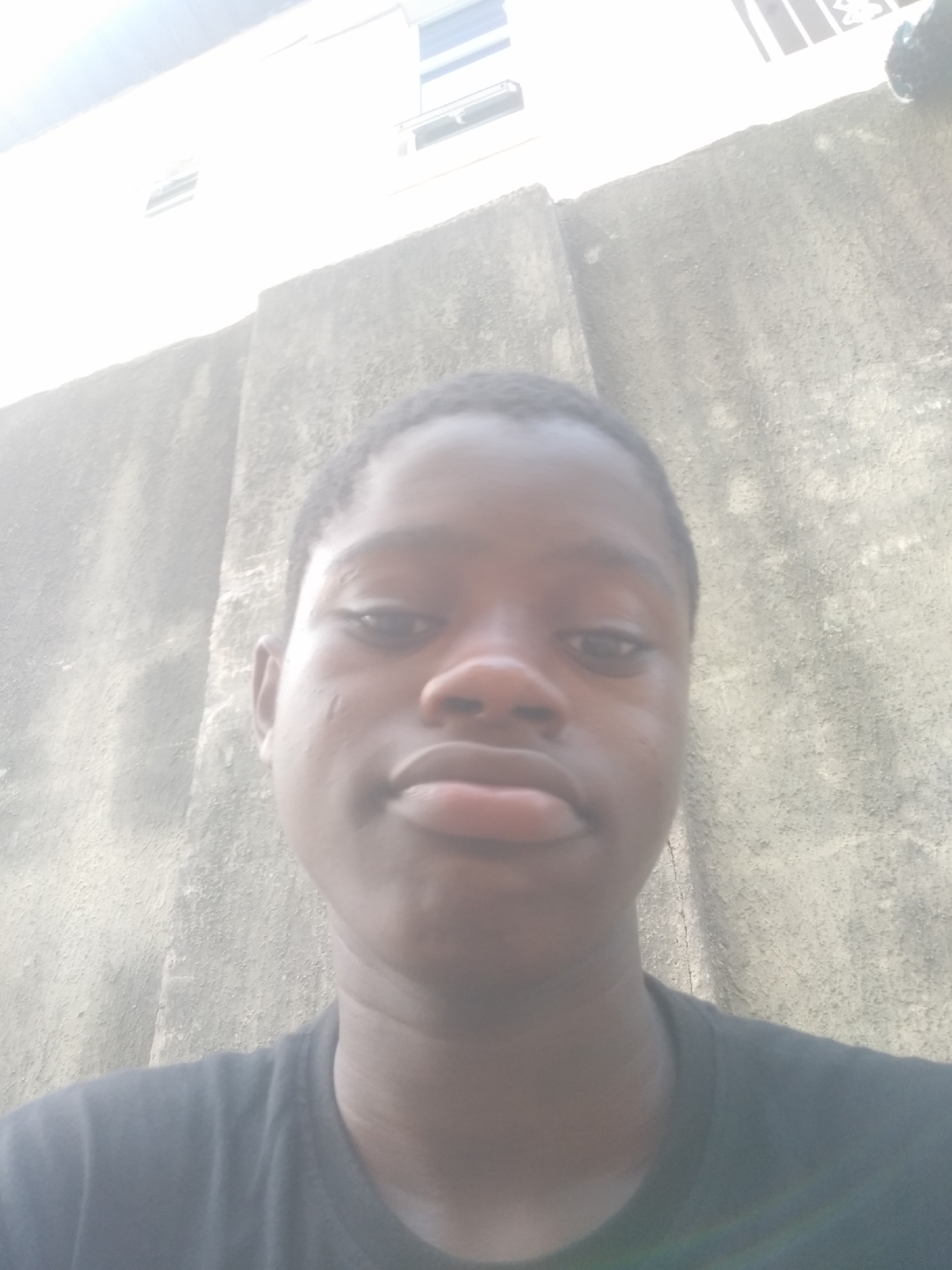 A person is standing in front of a wall. The person is dark-skinned and has short, dark hair. The person is wearing a black shirt. The wall is light gray and textured. Behind the wall is a white building with windows. The location is likely Shangisha, Nigeria.