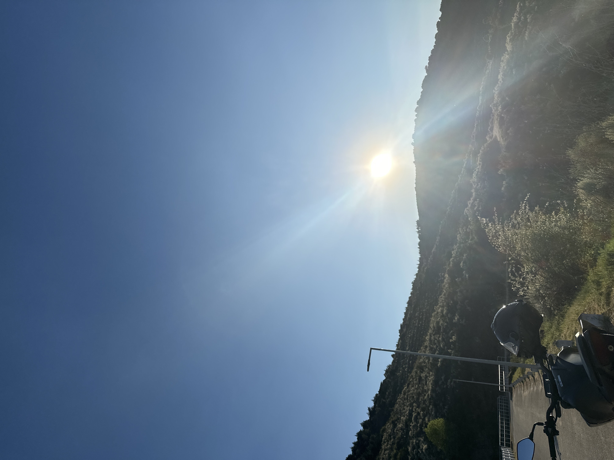 The image depicts a bright, sunny day in what appears to be a mountainous or hilly area. A motorcycle with a helmet resting on it is visible in the foreground, parked on an asphalt road. The background features a steep hillside covered in vegetation, possibly located outside the city of Mirandela in Portugal, but this cannot be confirmed without further information. An utility pole is visible near the hillside. The sun is high in the sky, causing glare and strong lighting conditions.
