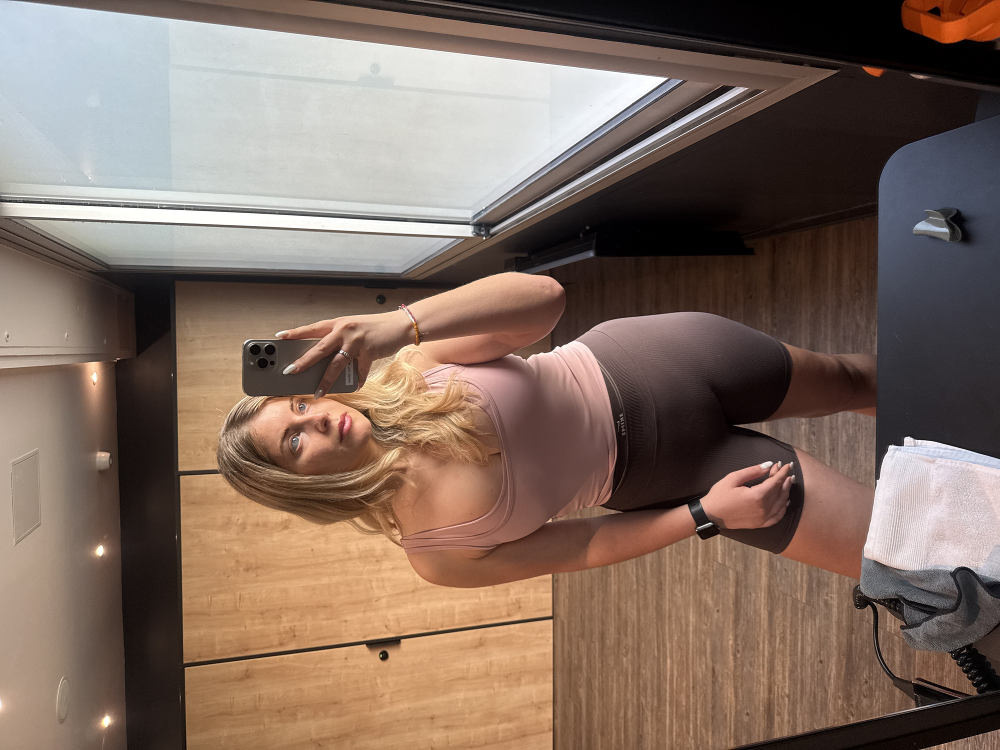 A full-body mirror selfie captures a woman standing indoors. She has blonde hair and is wearing a light pink tank top and dark brown ribbed athletic shorts. A black smart watch is visible on her left wrist, along with a ring on her left ring finger. A multicolored beaded bracelet is on her right wrist. She holds a silver-colored iPhone, which is reflected in the mirror as she takes the picture.
The setting appears to be a modern indoor space, possibly a gym or a contemporary changing room, characterized by light-colored wooden paneled walls. Several small, circular ceiling lights are visible on the left side of the reflection. A large window or skylight dominates the upper part of the image, showing a bright, diffused light from outside. To the right, a dark cabinet or piece of furniture is visible, along with a folded white and grey towel resting on a dark surface, and a black coiled cable. The text "SAMSAR" is faintly visible and mirrored on the waistband of her shorts. A sticker on the back of her phone, seen in the reflection, reads "MAKE THE MOST OF YOUR TIME". The woman looks directly at the camera with a neutral expression.