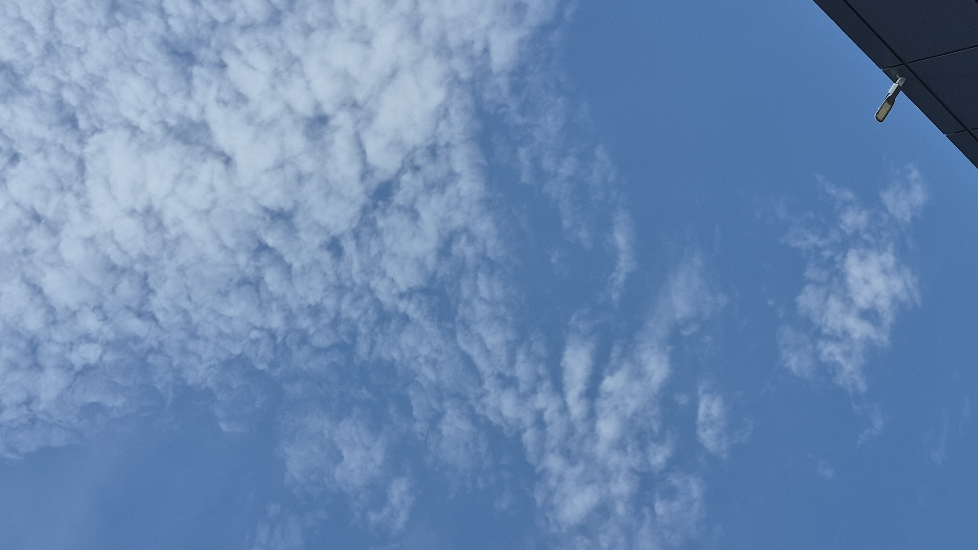 The image shows a portion of a dark gray building against a bright blue sky. The building occupies the lower right corner of the frame and has a modern, angular design. A single outdoor light fixture, likely an LED street lamp, is attached to the edge of the building. The sky is partly cloudy, with scattered, wispy white clouds filling the upper portion of the image. The overall scene is a typical urban outdoor environment on a clear day.