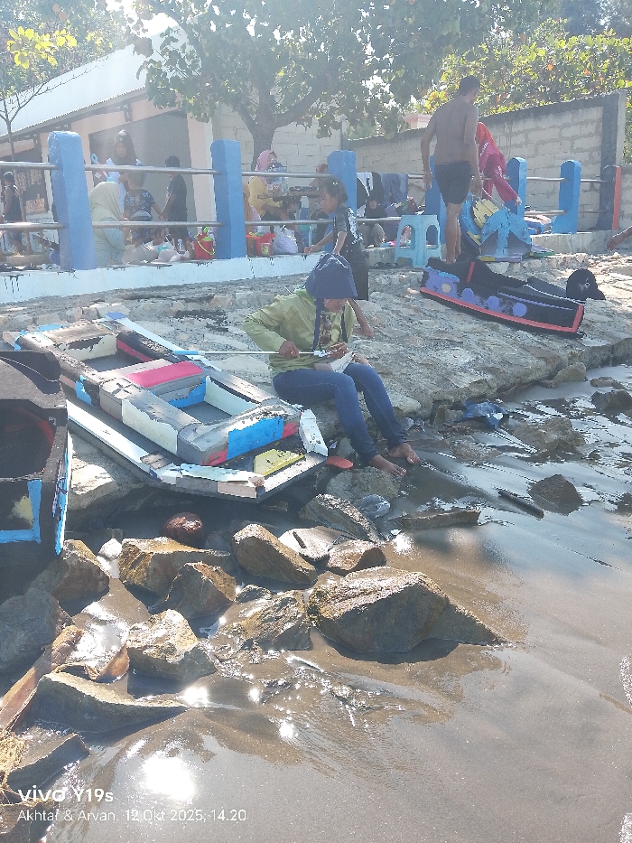 A person sits by a colorful, disassembled boat on a rocky beach, near the water's edge. The individual wears a hat and blue jeans, appearing focused on a task with a tool in hand. In the background, a group gathers by a blue and white railing, near a building. A figure stands near a toy resembling a train. The scene is sunlit, with glare reflecting on the wet sand and rocks.