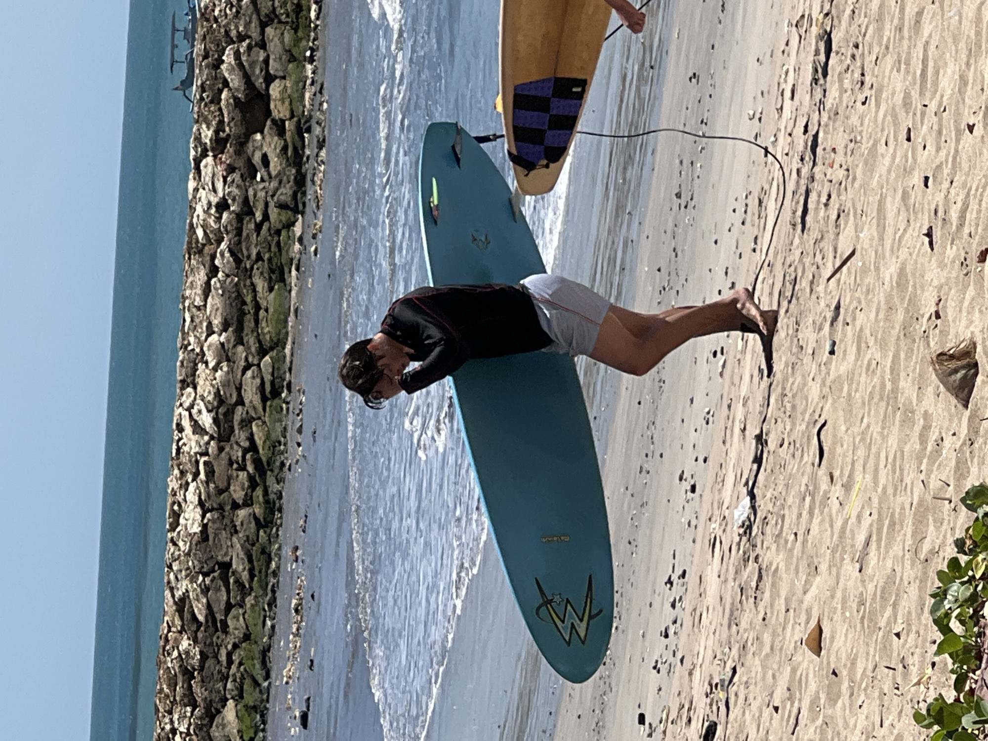 A young man, wearing a black rash guard and light-colored shorts, walks along the beach in Kuta, Indonesia, carrying a blue surfboard. He is facing left and appears to be adjusting his sunglasses or hair with his left hand. The surfboard has a logo on it that resembles the WWE logo. Another person, whose legs and a portion of a yellow surfboard with a black and purple checkered pattern are visible, is walking to the right of the man with the blue surfboard. They are both on a sandy beach with the ocean and a rocky breakwater in the background. The water is calm with small waves, and the sky is clear and blue.