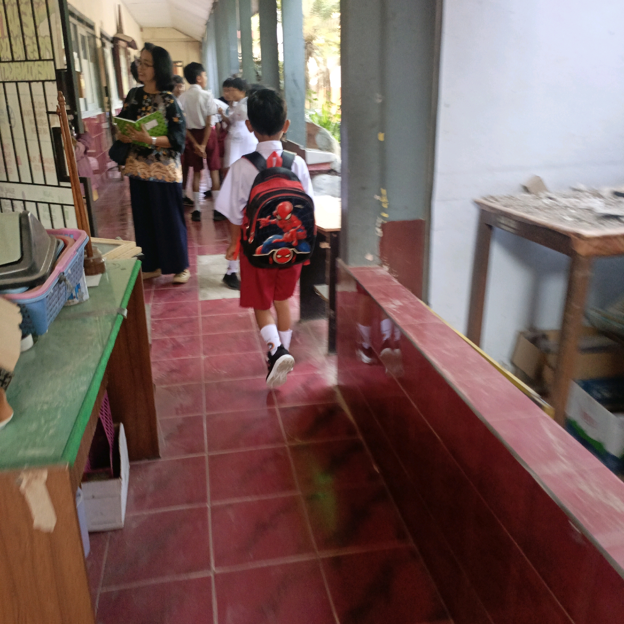 The image depicts a school hallway in Salatiga, Indonesia, with several children in uniform and an adult. On the left, a woman wearing a dark blue skirt and a patterned top stands holding a green book. She appears to be a teacher or guardian. In the center, a young boy walks away from the camera, wearing a white shirt, red shorts, white socks, and black shoes. He has a red and black backpack with a Spider-Man design. Other children in similar uniforms are visible in the background, some of whom appear to be talking to each other. The hallway has tiled floors with a reddish hue and a wooden railing along the right side. On the left, there is a table with a blue plastic basket containing what looks like metal trays, and a wooden cabinet. The environment appears to be an indoor school setting, possibly during a break or transition between classes.
