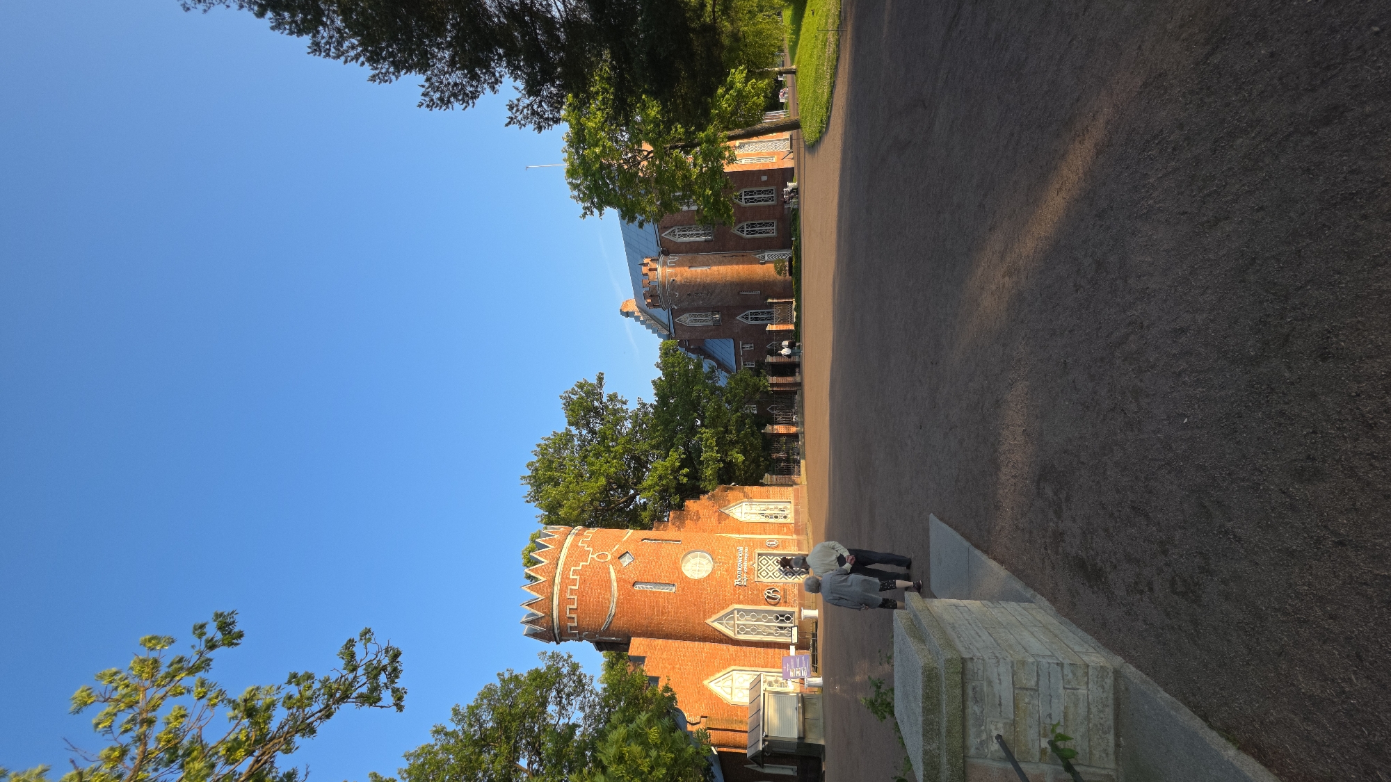 The image depicts an outdoor scene with a clear blue sky and lush green trees framing the view. In the foreground, a low, red brick building with crenellated battlements and arched windows stands prominently. A gravel path runs alongside the building, leading towards a larger, darker red brick structure in the background. This larger building also features multiple arched windows and appears to be a significant architectural landmark.
Two people, dressed in casual attire, are visible near the entrance of the smaller building, appearing to converse or observe something. The warm sunlight suggests it is either early morning or late afternoon, casting a gentle glow on the brickwork. The overall atmosphere is serene and picturesque, typical of a park or historical estate.
There is some text visible on a sign mounted on the smaller building, but it is too blurry to be read clearly. The image is captured from a slightly tilted angle, adding a dynamic feel to the composition.