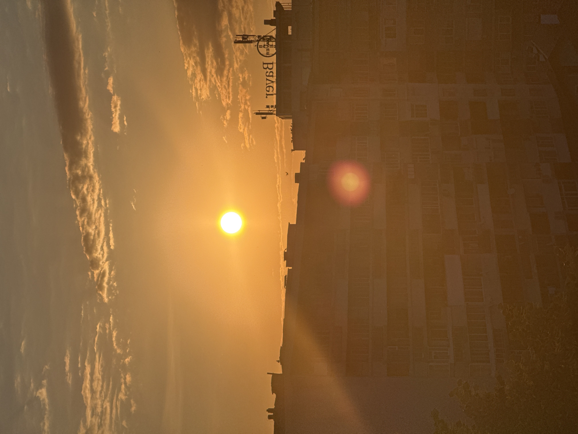 The image shows a sunset over the city of Dobrich, Bulgaria. The sun is a bright, glowing orb in the center of the sky, casting a warm, orange light over the scene. Silhouetted buildings with numerous windows rise from the foreground, their details softened by the haze of the sunset. Some buildings have antennas on their roofs, and one has a large sign with reversed lettering that appears to read "BAKER". A few birds are visible in the sky. The overall impression is one of urban tranquility as the day ends.