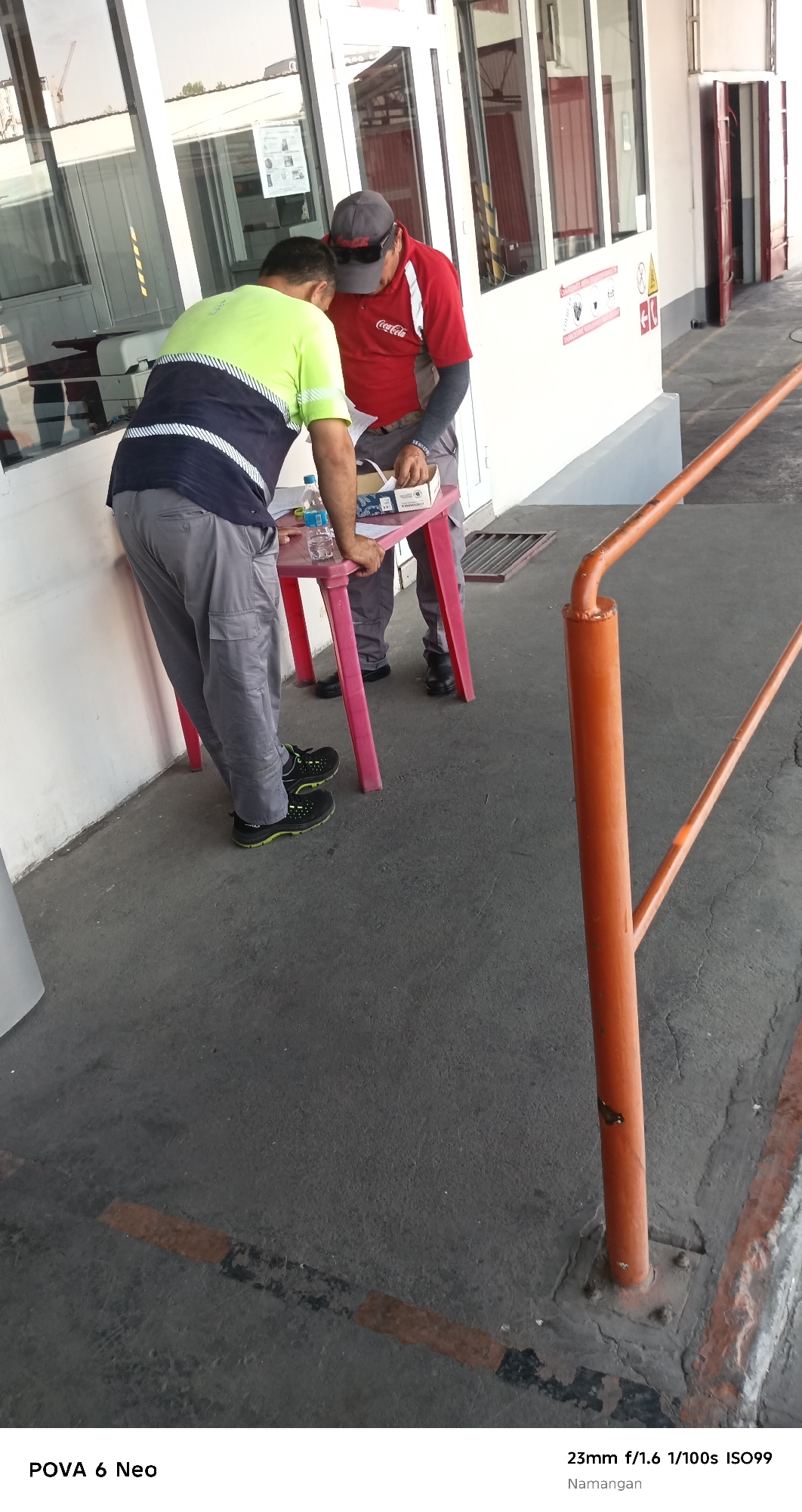 Two men are standing near a pink table outside a building with large windows. One man, wearing a lime green and navy shirt and gray pants, is looking down at the table. The other man, wearing a red shirt with a Coca-Cola logo and a grey baseball cap, is also looking at the table, and appears to be handing something to the first man. On the table are a bottle of water, a box, and some papers. The ground is concrete, and an orange metal barrier is visible on the right side of the image. The scene is set in Namangan City, Uzbekistan.