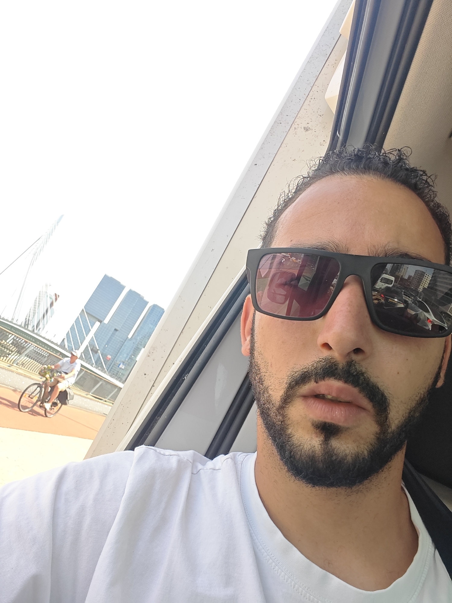 This media file is a selfie taken from inside a vehicle, showing a man with dark, curly hair and a beard, wearing sunglasses and a white t-shirt. The sunglasses reflect a busy street scene with buildings and a vehicle. In the background, through the window, a person is seen riding a bicycle on a bridge. Tall modern buildings and a suspension bridge are visible, suggesting an urban setting. The bright sky indicates it is daytime, likely a sunny day. The overall mood appears casual and relaxed. The reflection in the sunglasses offers a glimpse of the external environment, which includes a white van and other vehicles. The architecture of the buildings and the style of the bridge are characteristic of Rotterdam, Netherlands.