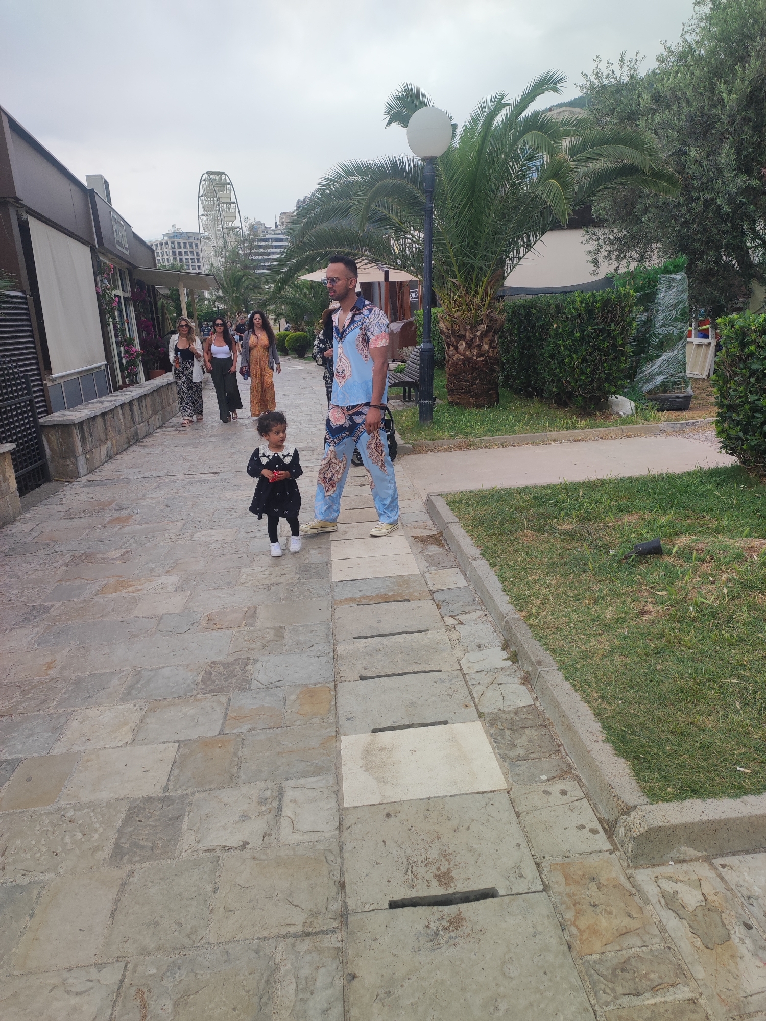 This image captures an outdoor scene in Budva, Montenegro, on an overcast day, depicting a paved promenade or walkway flanked by buildings and greenery.
In the foreground, a man and a small child are walking. The man wears a distinctive light blue shirt and matching trousers with an intricate pattern, sunglasses, and has a small bag slung over his shoulder. He is seen walking towards the right. To his left, a young child, possibly a toddler, walks in a black dress with a white collar and white shoes, holding a small red object.
Further down the paved path, several women are strolling. One woman wears a white top and patterned trousers, another a white tank top and dark trousers, and a third a long orange dress. On the left side of the path are low-rise buildings, possibly cafes or shops, with some plants and flowers visible. A sign partially displaying the word "CAPRI" is visible on one of these buildings.
To the right of the walkway, there is a strip of green grass and neatly trimmed hedges, interspersed with tall palm trees and other lush foliage. A street lamp with a spherical light fixture stands near a palm tree, and a partial view of a bench is visible behind it. In the grass area, a white cat can be seen lying down. In the background, against a cloudy sky, a large ferris wheel is prominently visible above the city buildings, indicating a vibrant tourist area. The pavement consists of irregular grey stone slabs.