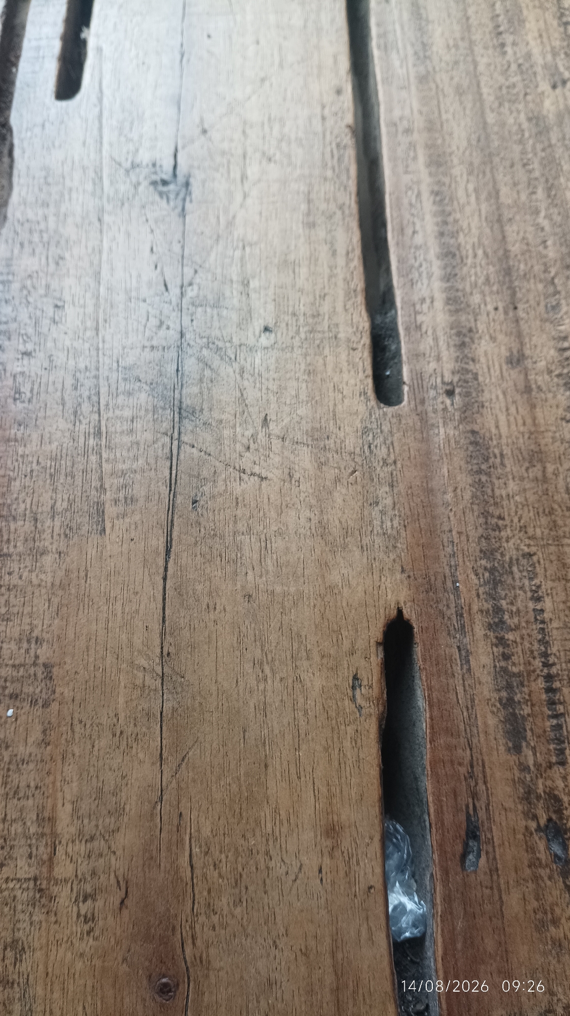 A close-up, overhead shot shows the surface of a wooden table. The wood has a warm, brown tone with visible grain and a rough texture, featuring several long, dark gaps running vertically down the image. Some of these gaps appear to be carved or naturally occurring. A small amount of plastic debris is visible within one of the gaps. The date and time "14/08/2026 09:26" are imprinted in the lower right corner of the image. The setting appears to be outdoors or in an area with natural light, given the texture and color variations in the wood. There are no people or animals visible in the image, and the focus is solely on the material and texture of the wooden surface.