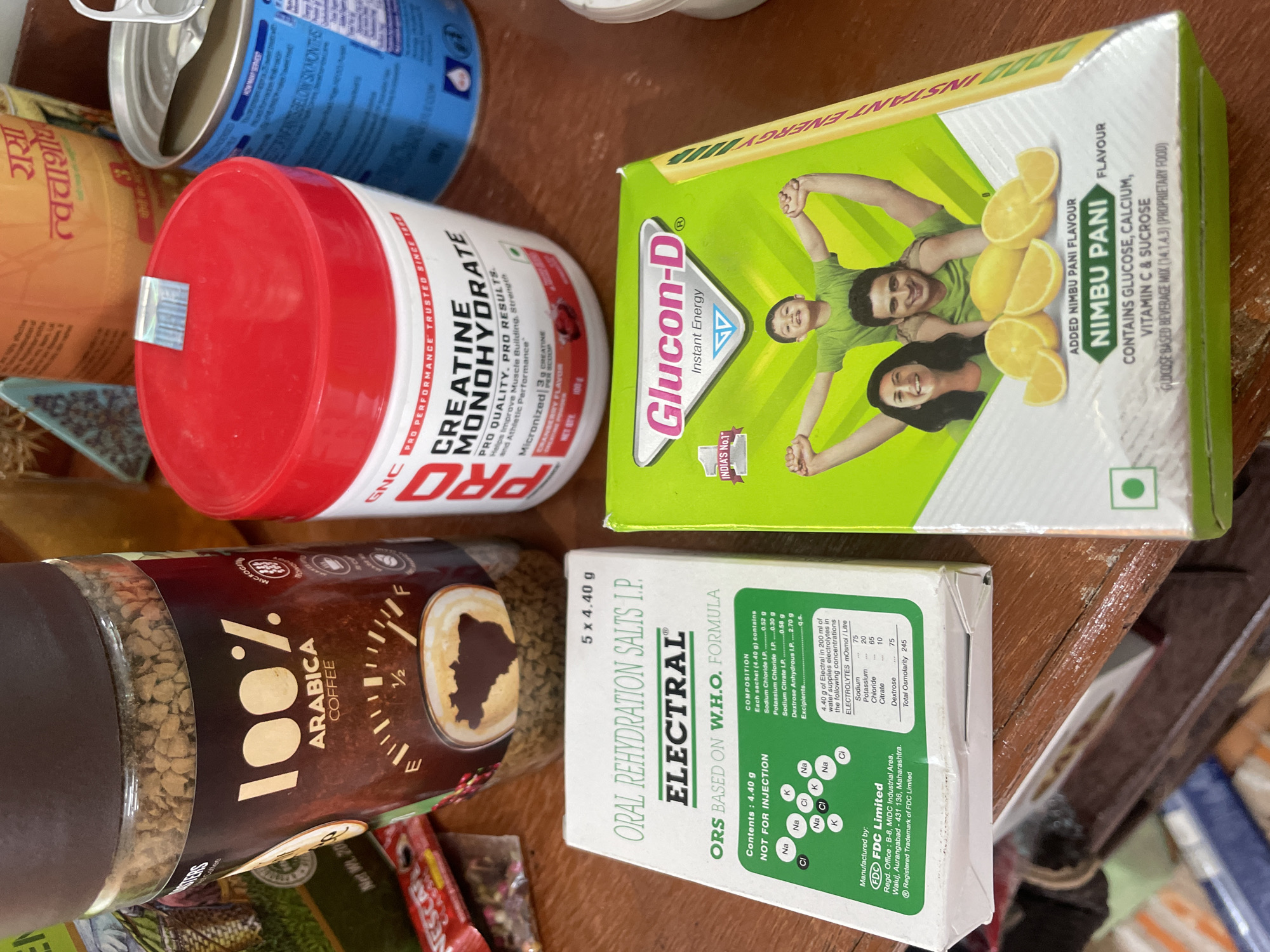 The image shows a collection of health and fitness products arranged on a wooden surface. In the foreground, on the left, is a white and green box labeled "ELECTRAL ORAL REHYDRATION SALTS I.P. ORS BASED ON W.H.O. FORMULA". Next to it, on the right, is a bright green box for "Glucon-D Instant Energy" featuring images of a family and lemons. Behind these boxes, to the left, is a glass jar of "100% ARABICA COFFEE". To the right of the coffee jar is a white plastic container with a red lid, labeled "CREATINE MONOHYDRATE". A blue and silver can is visible to the far right, partially obscured. The background includes parts of other packaged goods, some with Hindi text. The lighting suggests an indoor setting. The overall scene depicts a variety of consumable items.