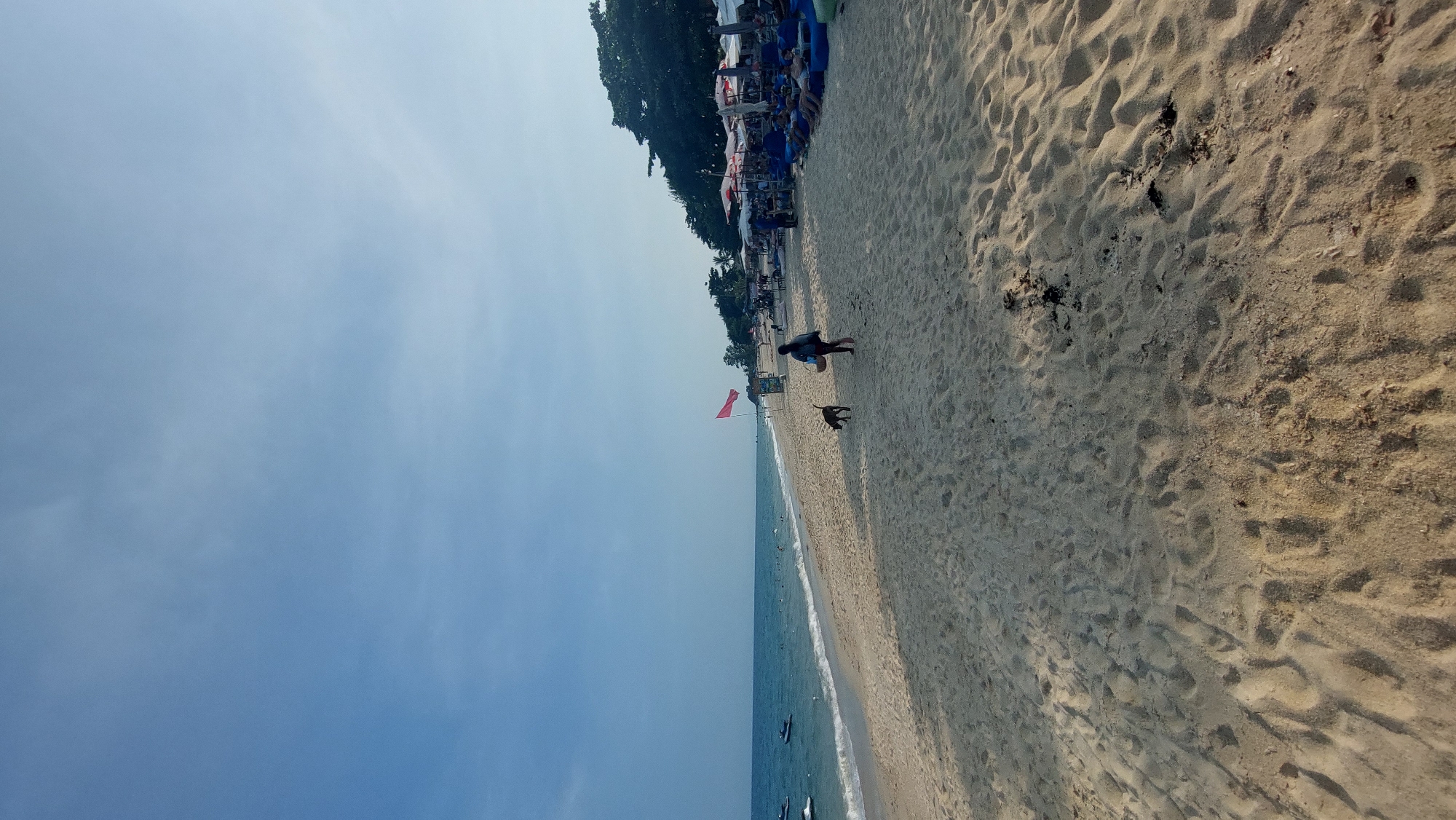 This image captures a serene, sunny day at a sandy beach in Baan Lamai, Thailand. The scene features a clear blue sky with subtle, wispy clouds in the upper portion, transitioning to a calm, blue ocean on the left side of the frame. A wide expanse of light-colored sand stretches across the right, showing distinct ripple patterns and scattered darker debris.
On the sandy shore, a person in dark clothing stands near the water's edge, accompanied by a small, dark-furred dog. Further down the beach, closer to the water, several individuals are visible in the ocean. A prominent red flag is planted in the sand, serving as a noticeable marker. Along the back of the beach, a dense line of dark green trees provides shade, beneath which numerous blue and white beach chairs and umbrellas are arranged, indicating a recreational area. Several small boats are seen in the ocean. The bright sunlight casts strong shadows on the sand, suggesting a midday or early afternoon setting. No discernible text is visible in the image.